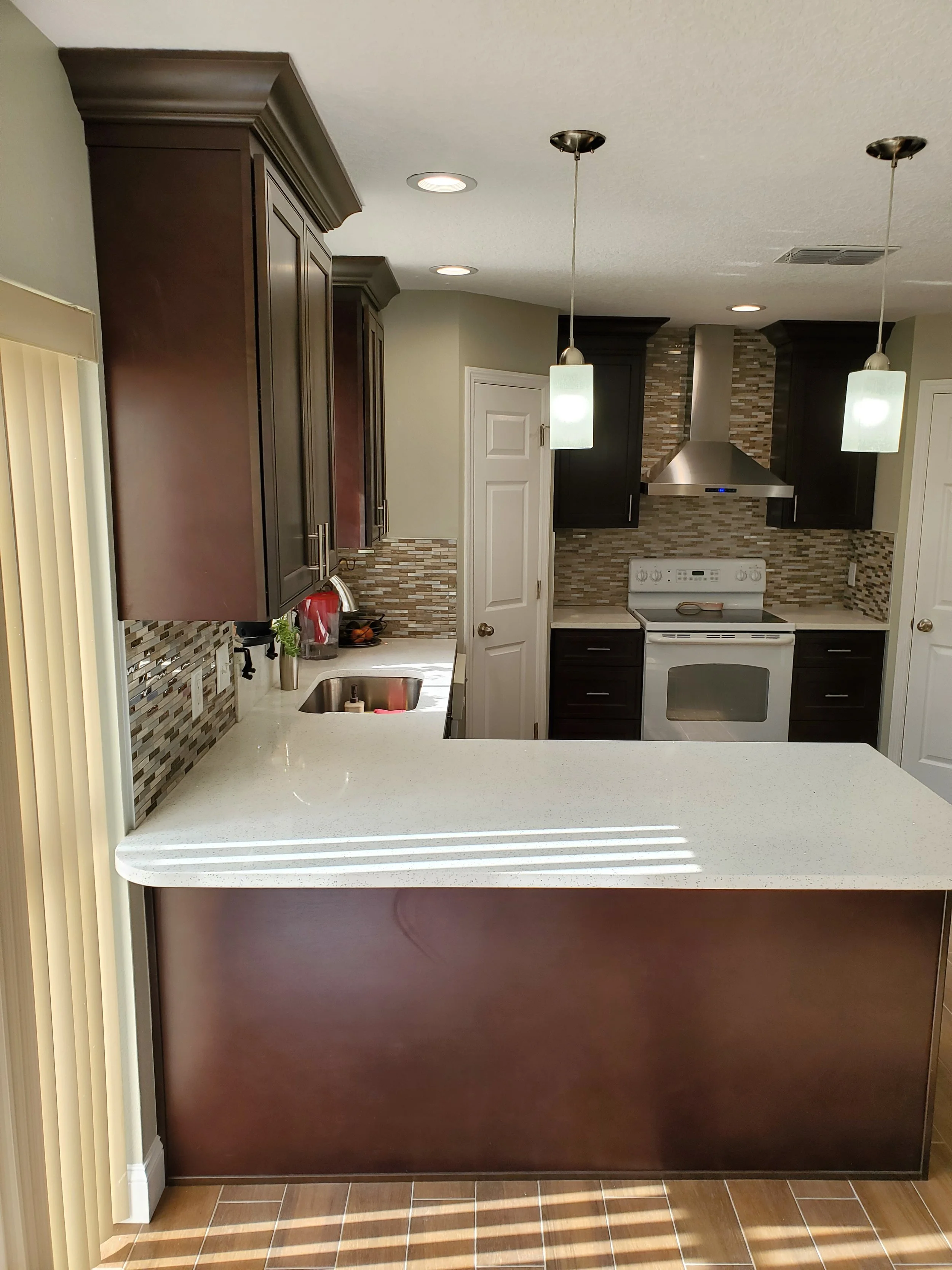 Modern kitchen with dark wood cabinets, beige countertops, mosaic tile backsplash, stainless steel range hood, white oven, and pendant lighting with frosted glass shades.