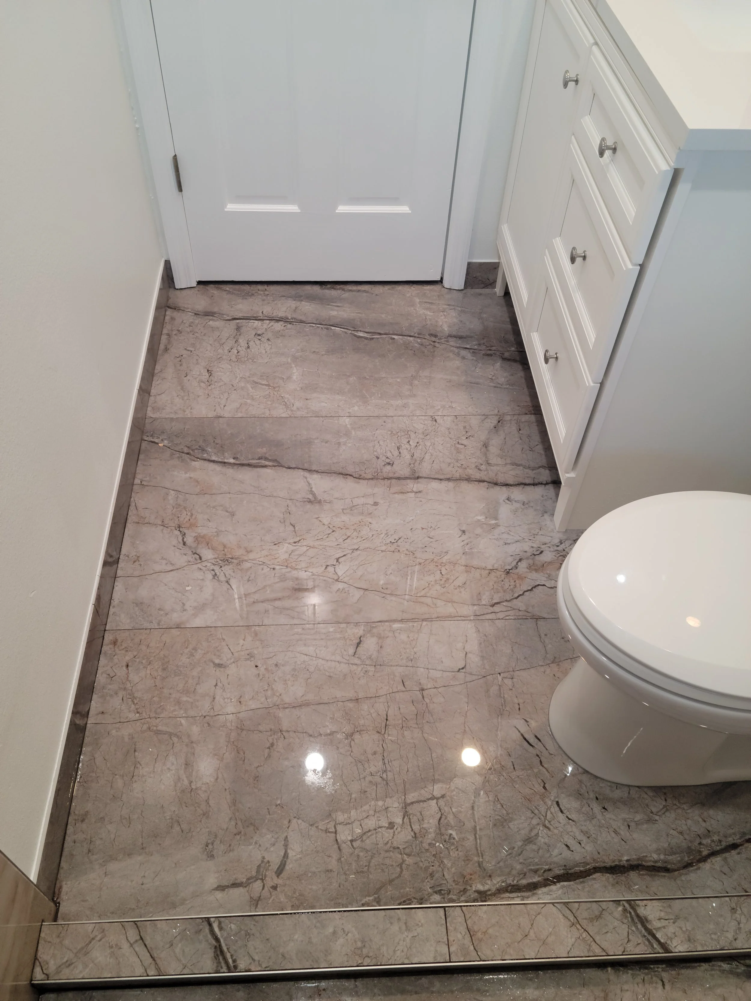 Close-up of a bathroom with polished marble flooring, a white toilet, white cabinetry, and a closed white door.