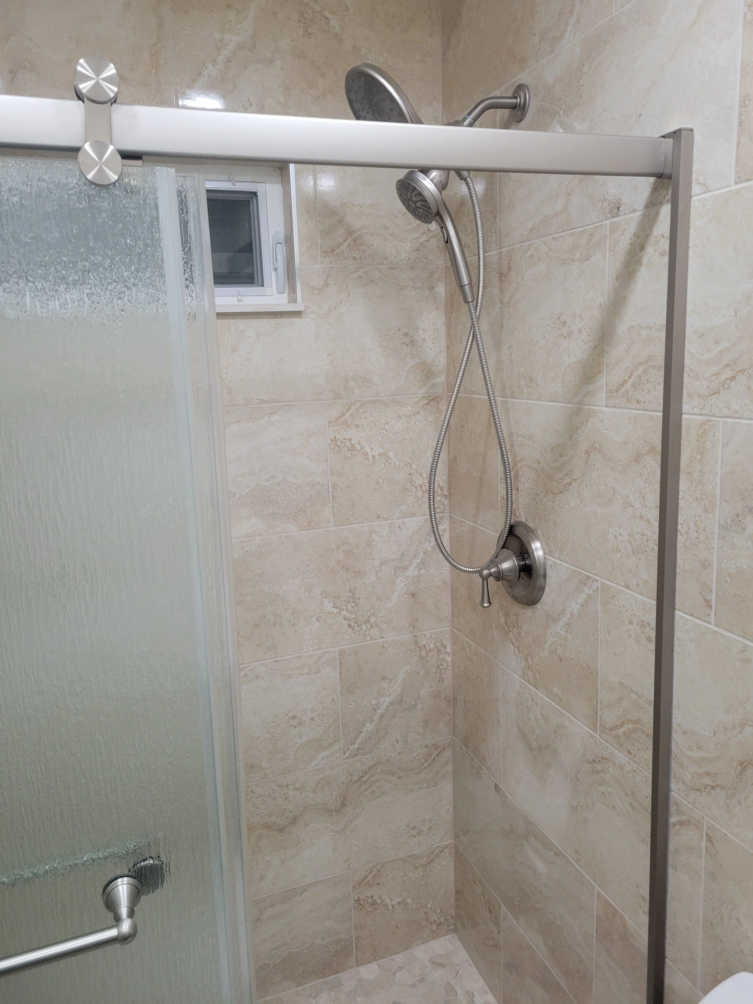 Shower with beige tiled walls, metal hand-held showerhead, and a glass door.