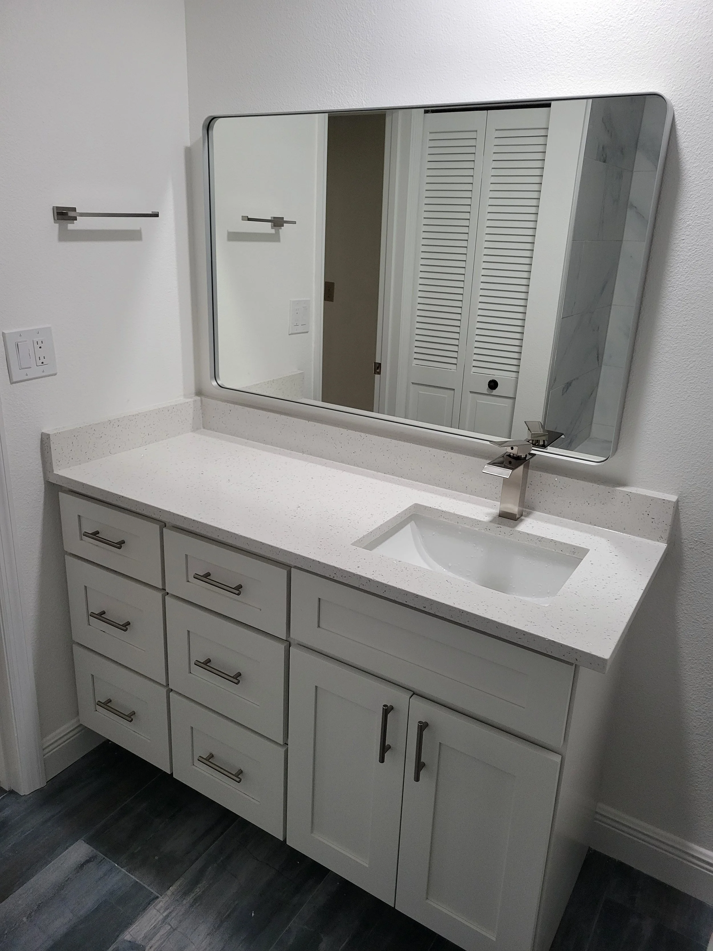 Bathroom vanity with white cabinet, speckled white countertop, small rectangular sink, large mirror, and modern faucet, reflected door with white louvered closet doors.