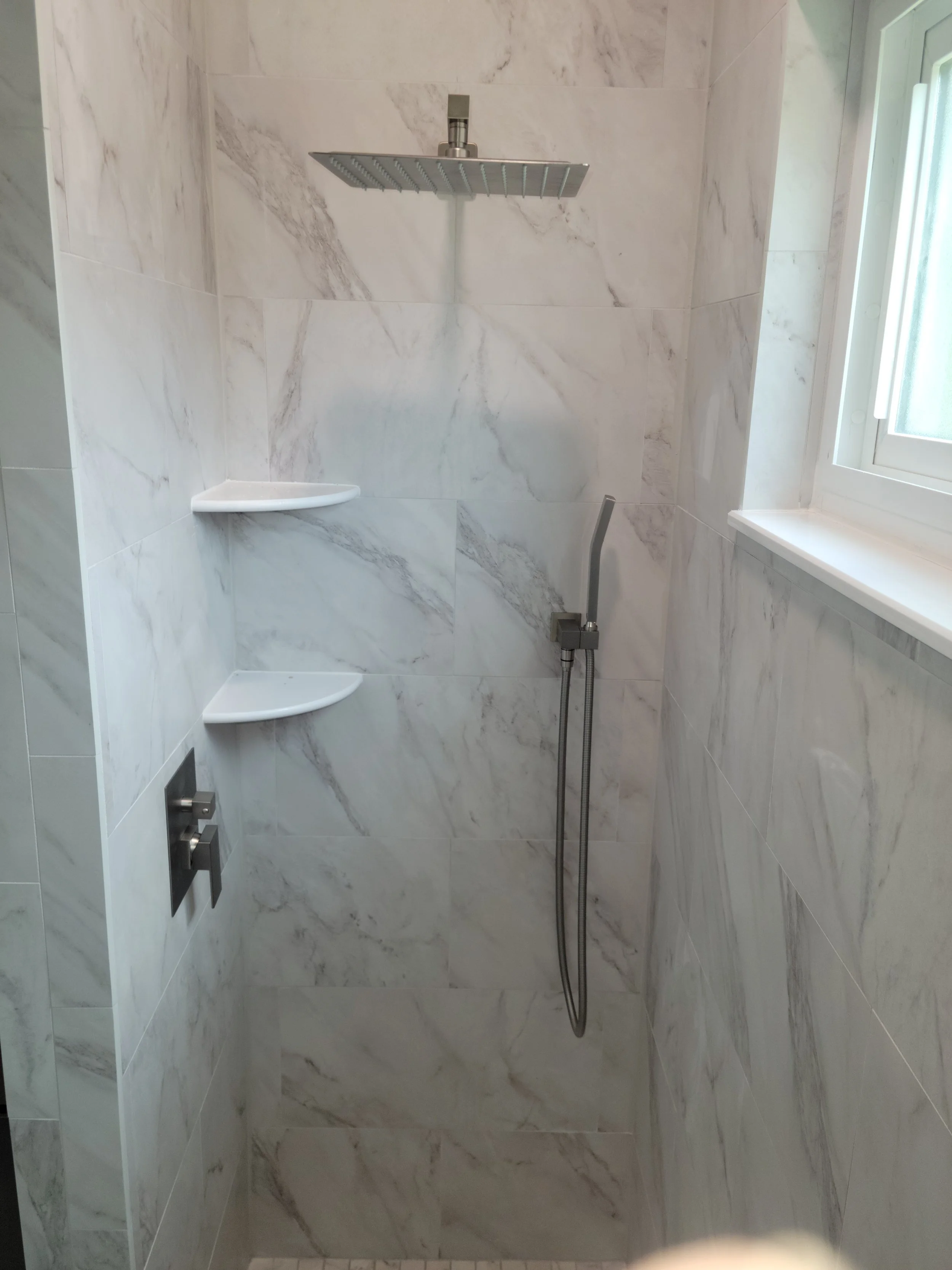 Shower area with marble tiles, a ceiling-mounted rain shower head, a hand-held shower wand, a small corner shelf, and a window.