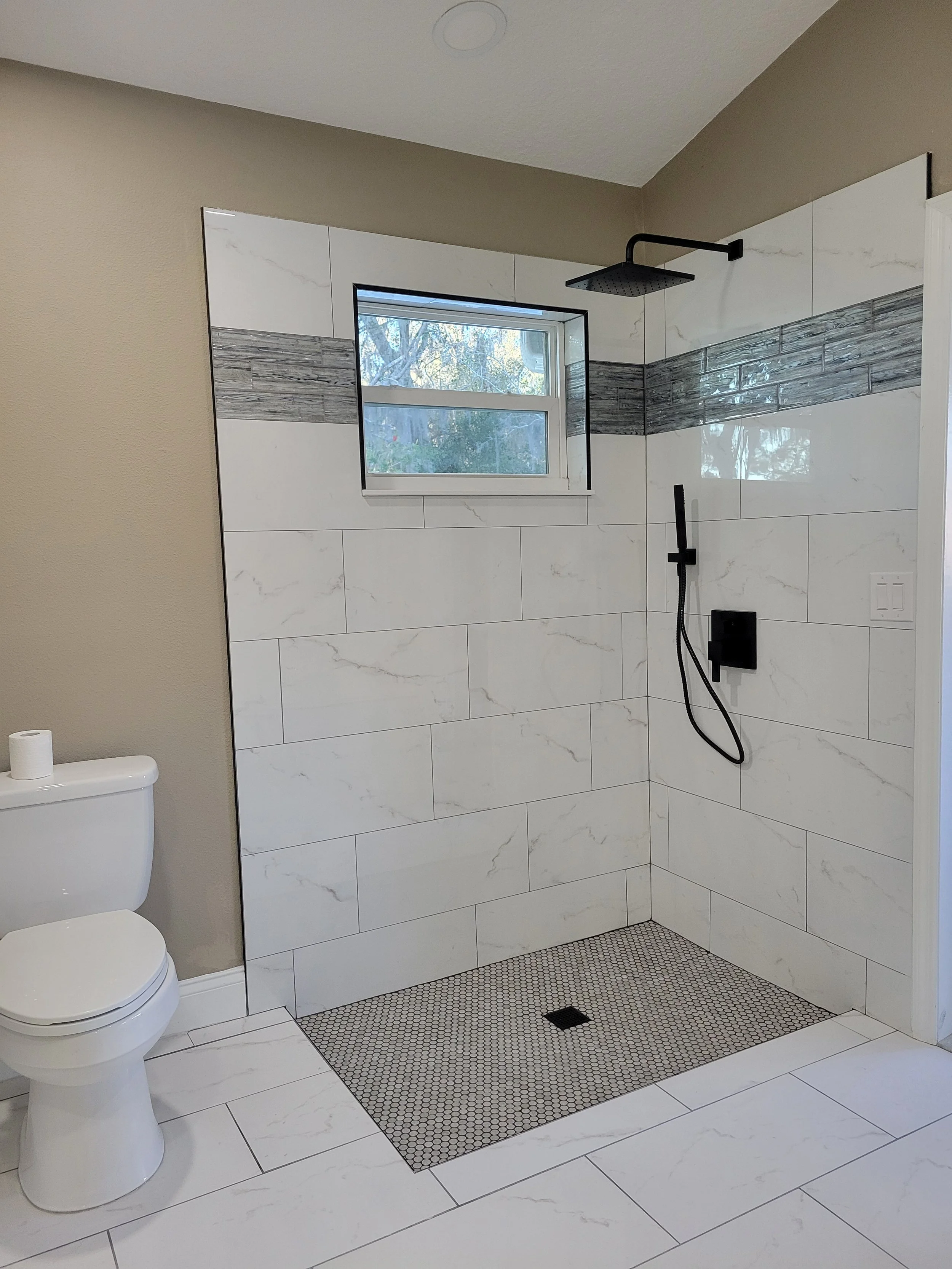 Modern bathroom with a walk-in shower featuring white and gray tiles, a small window, and a black showerhead. A toilet is visible on the left. The shower has a black handheld shower and a ceiling-mounted black rain showerhead.