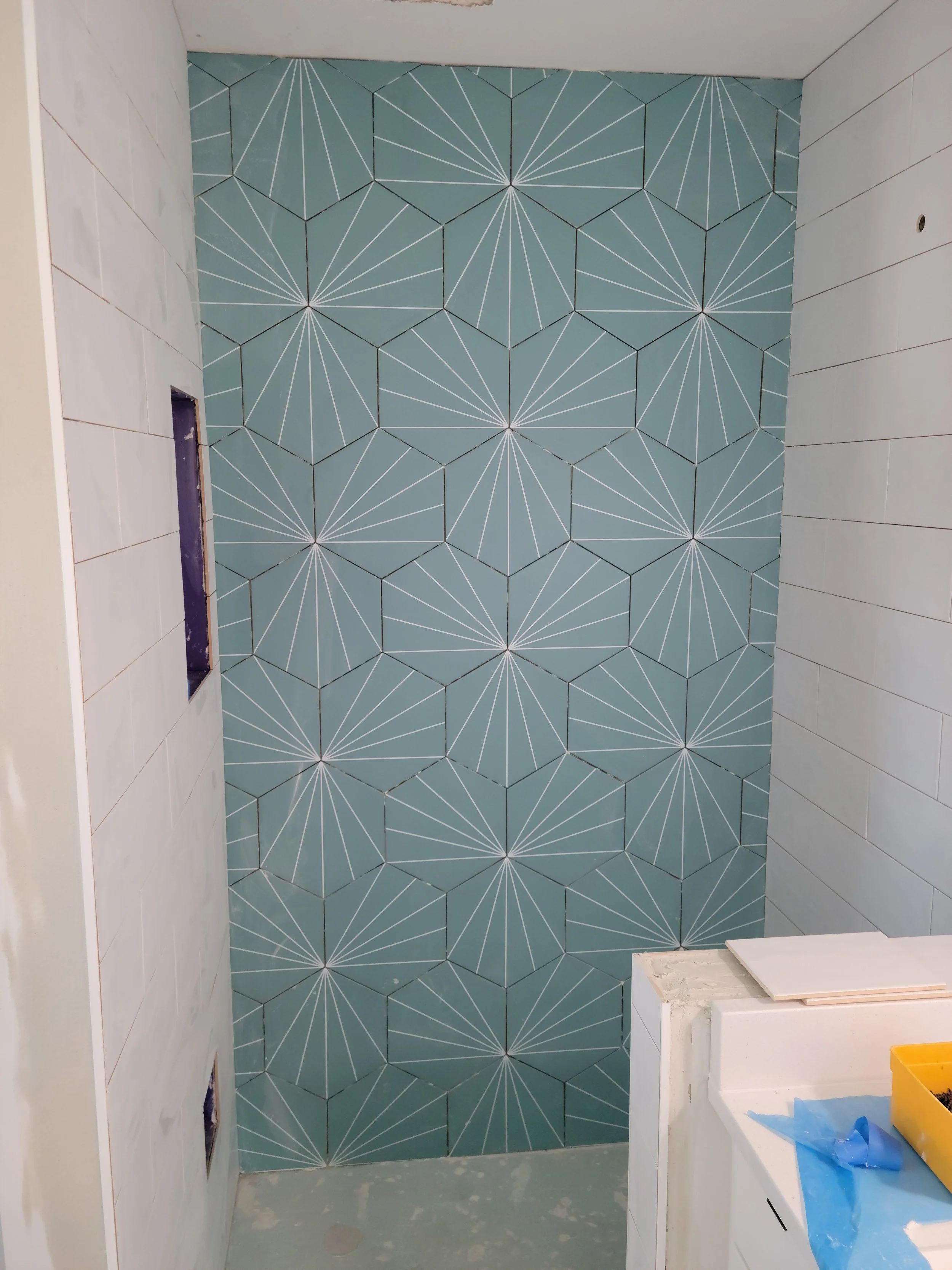 A bathroom shower area with a wall tiled in geometric blue tiles with white lines creating a starburst pattern, surrounded by white tiled walls, and some construction materials visible in the foreground.