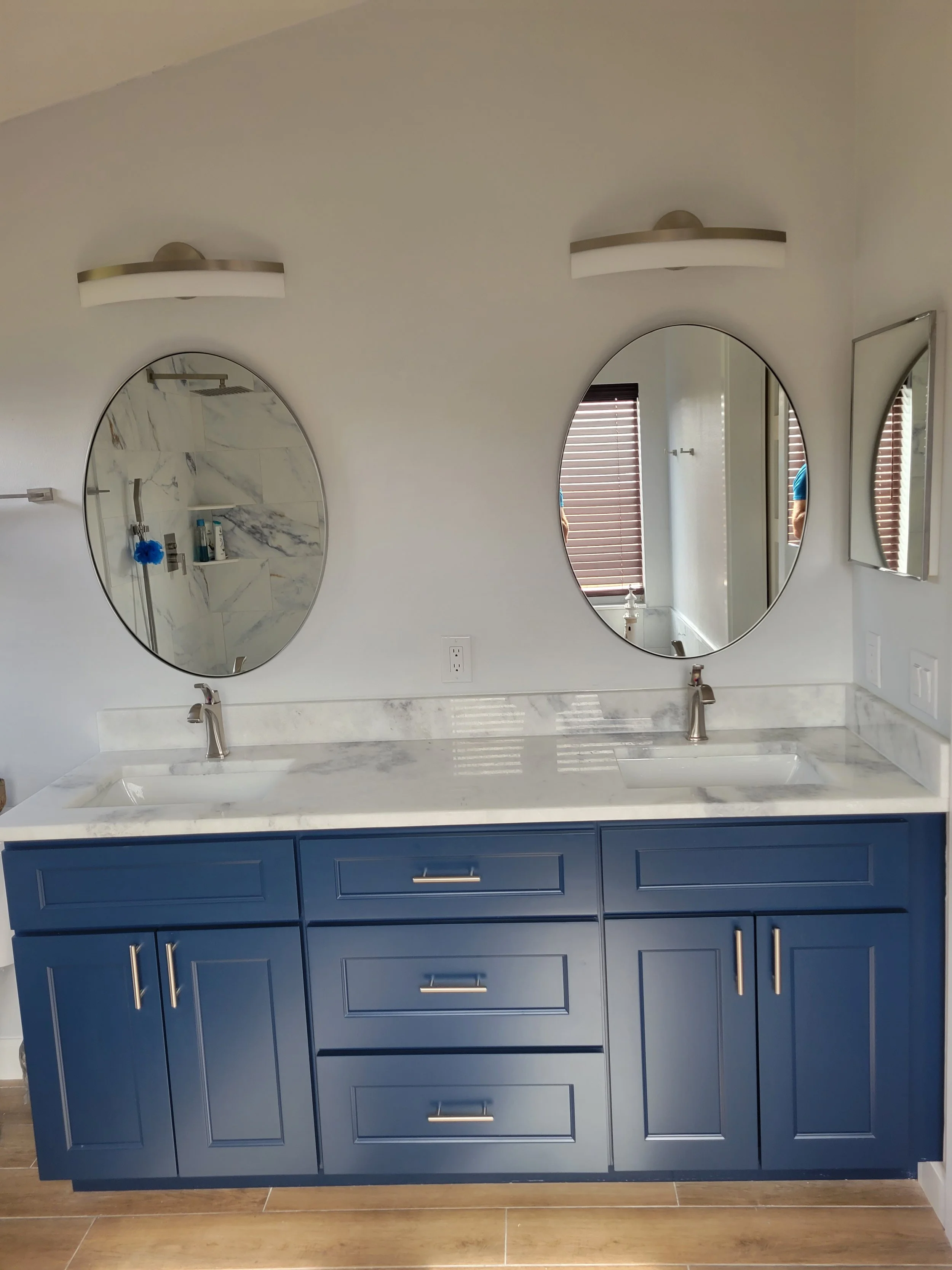Double vanity bathroom with blue cabinets, white marble countertop, two oval mirrors, and modern light fixtures