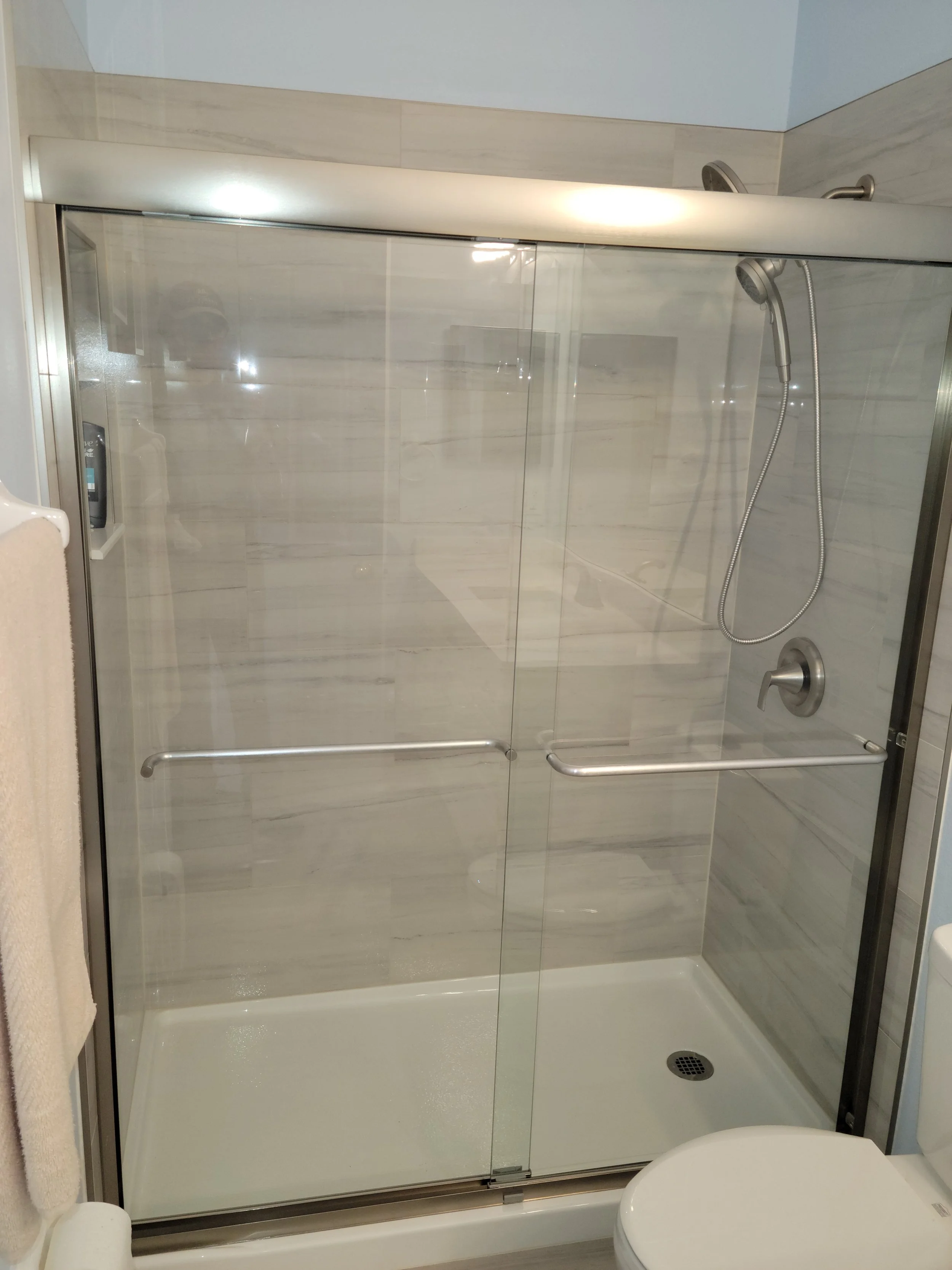 A modern walk-in shower with sliding glass doors, a handheld showerhead, a wall-mounted control, and a built-in niche for toiletries, in a bathroom with light-colored tile walls.