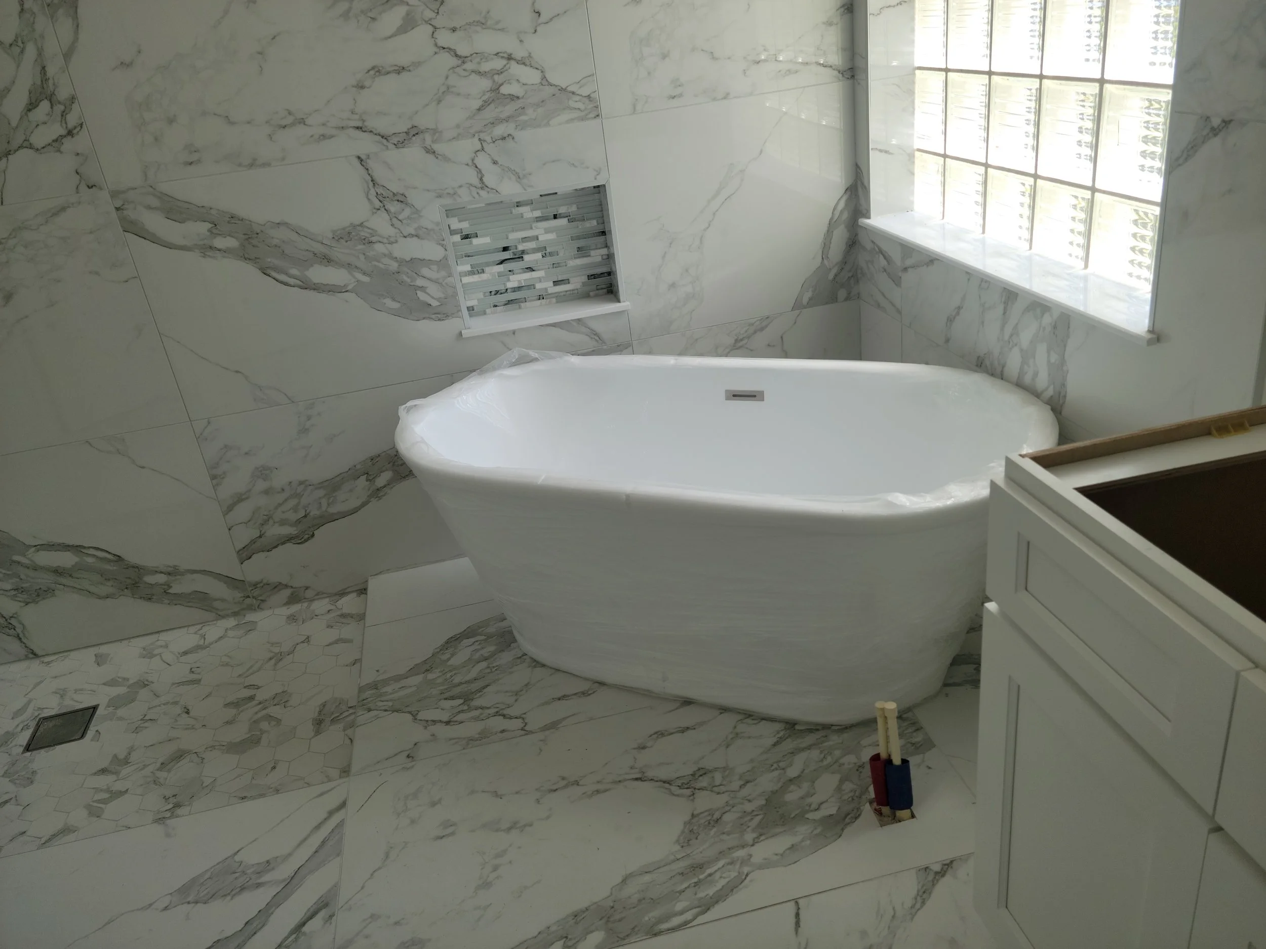 A bathroom with marble tiles on the walls and floor, featuring a freestanding bathtub next to a window, with a small vent in the wall and a white cabinet nearby.