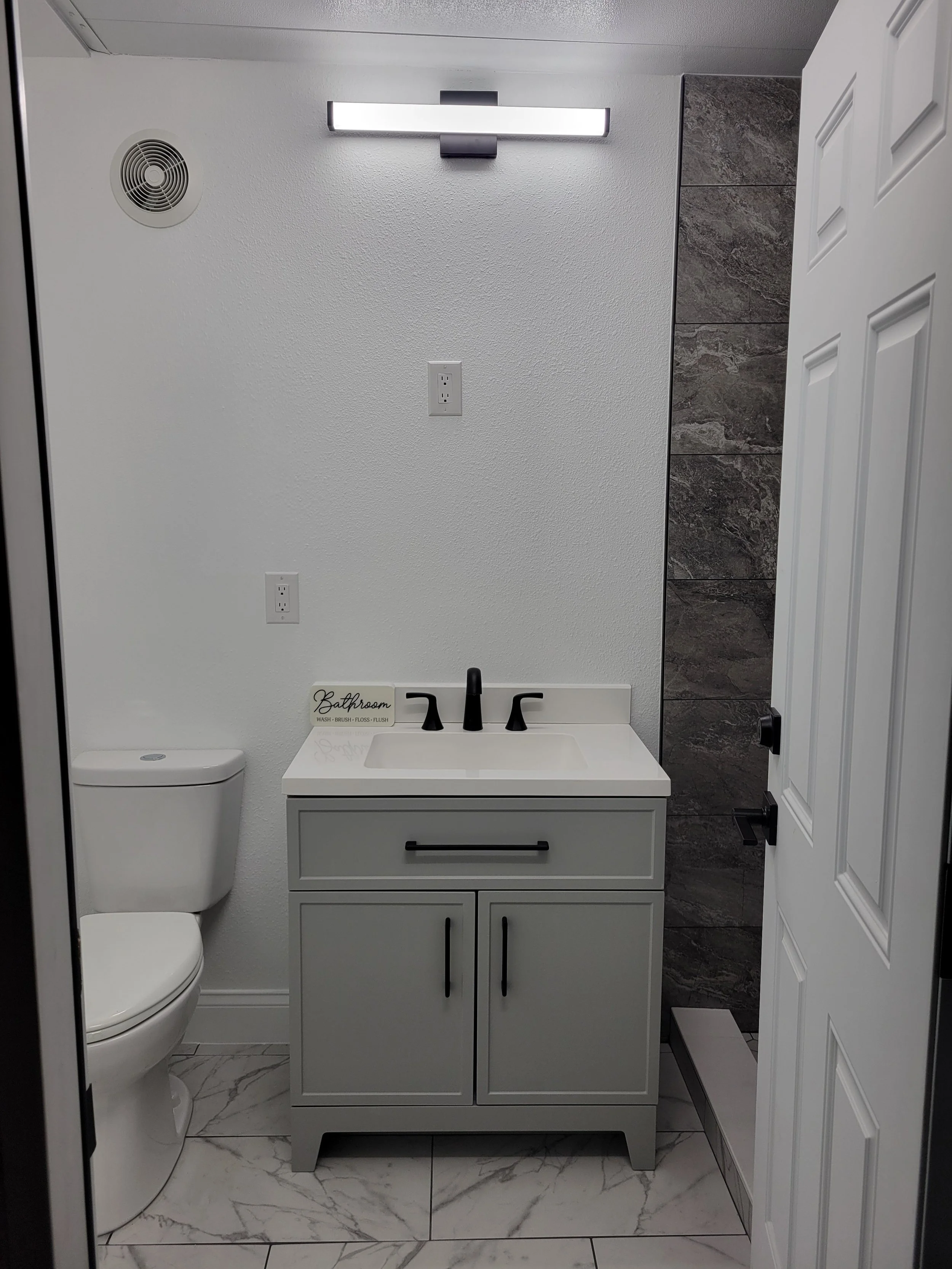 A small modern bathroom with a white toilet, a white vanity with a black faucet, white walls, a horizontal light fixture, a gray tiled accent wall, and a white door.