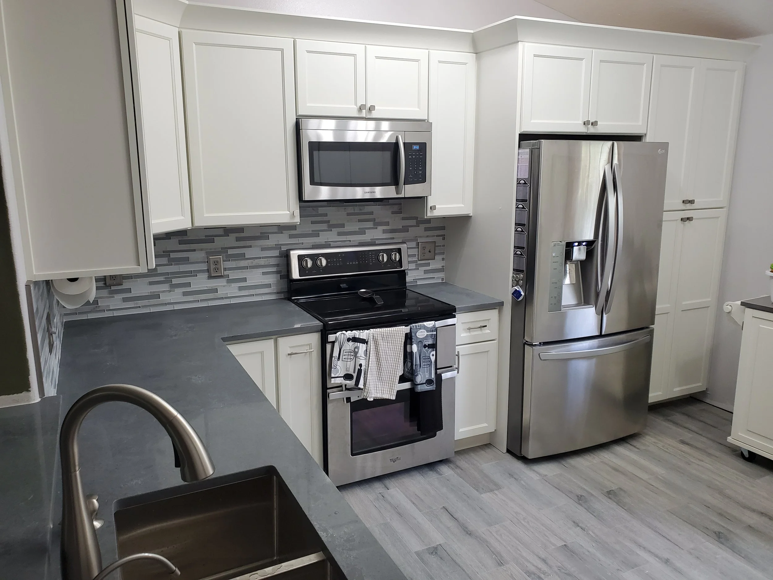 Modern kitchen with white cabinets, stainless steel appliances including a microwave, refrigerator, and oven, gray backsplash, and gray countertops with a sink in the foreground.