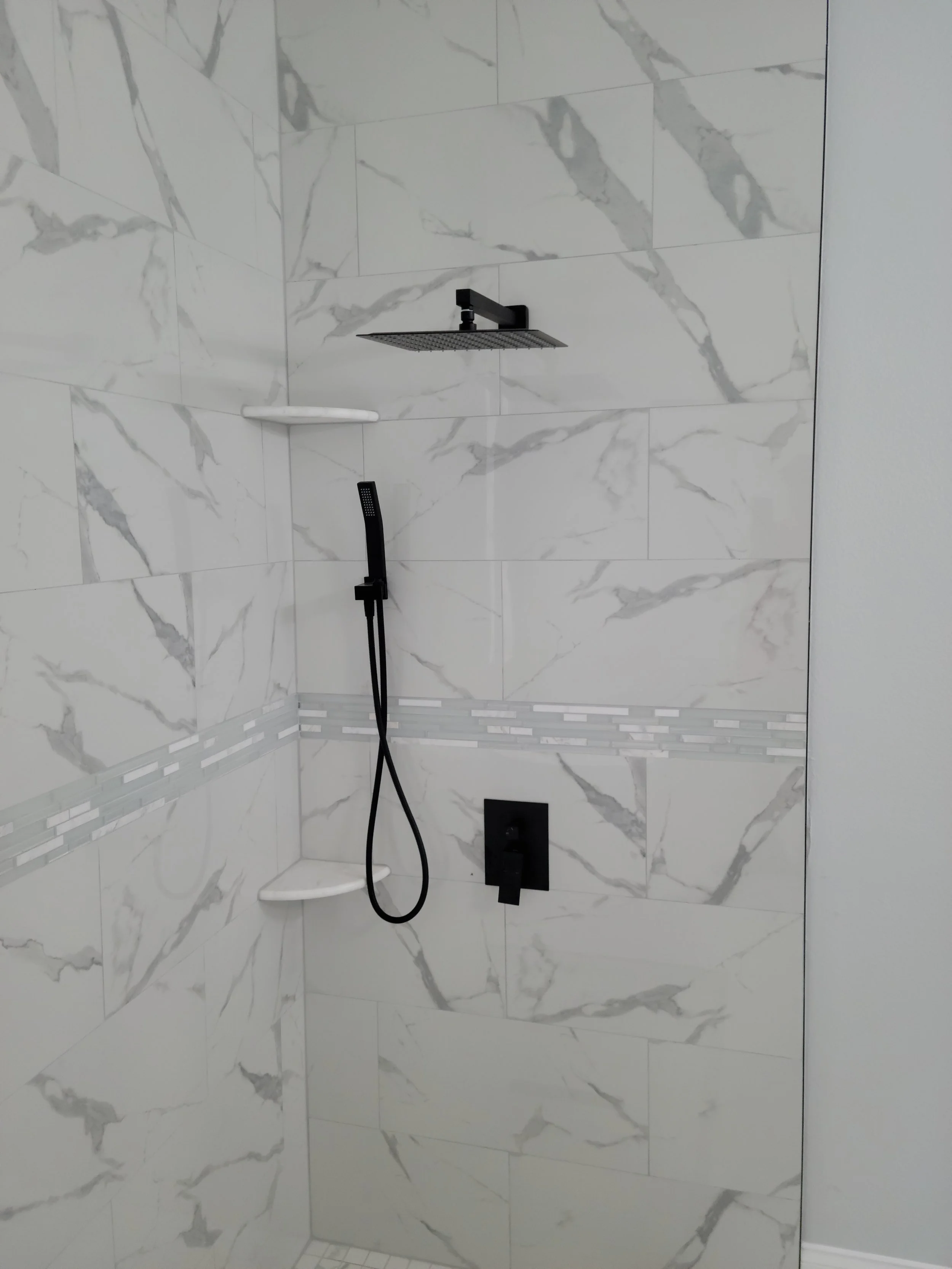 Modern walk-in shower with marble tile walls, a black rainfall showerhead, a black handheld shower, and corner shelves.