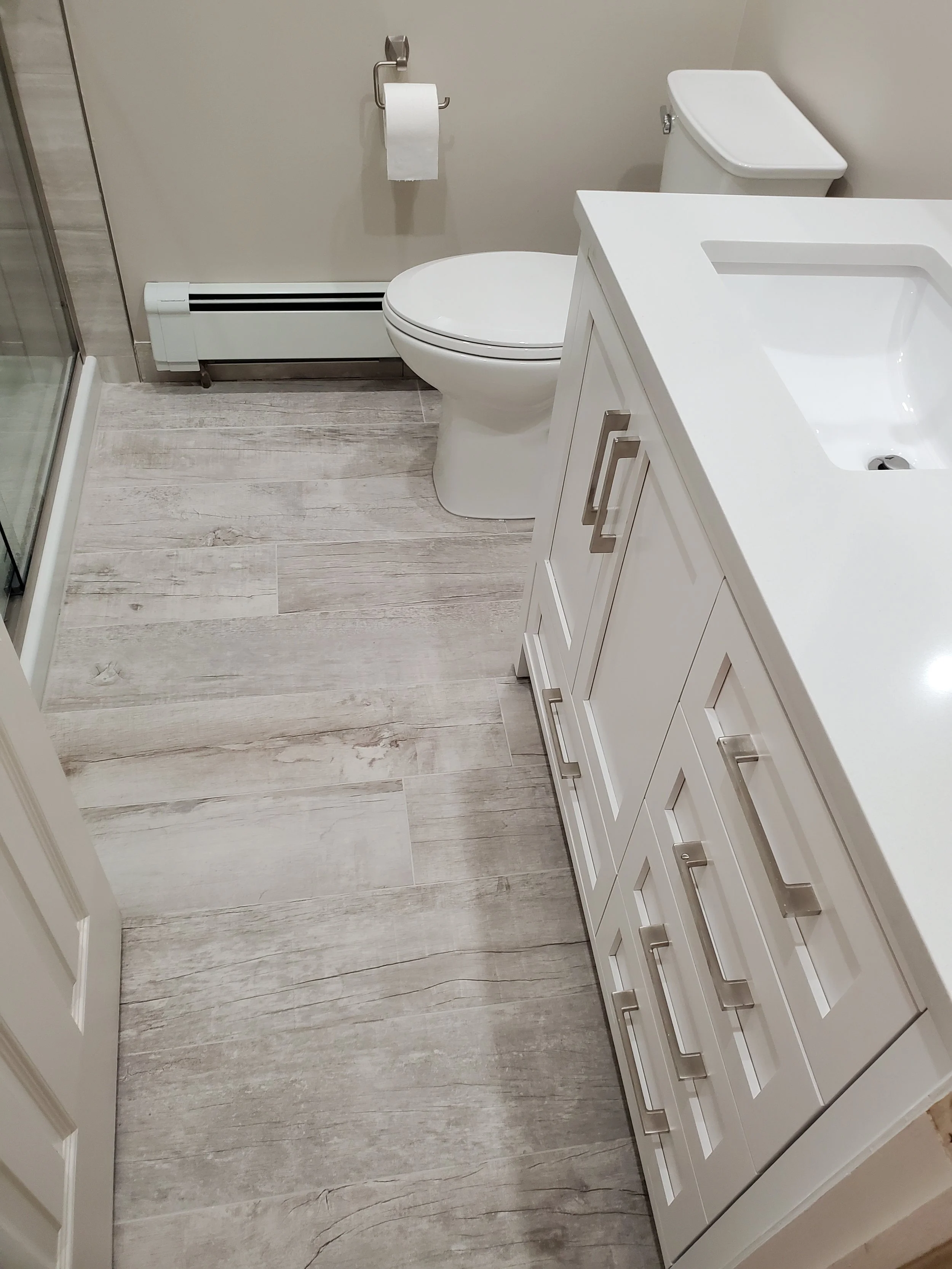 A small bathroom featuring a white toilet, a white vanity with a sink, a mirror, a toilet paper holder with paper, and a glass shower enclosure with a metal frame. The floor is covered with light wood-look tiles.