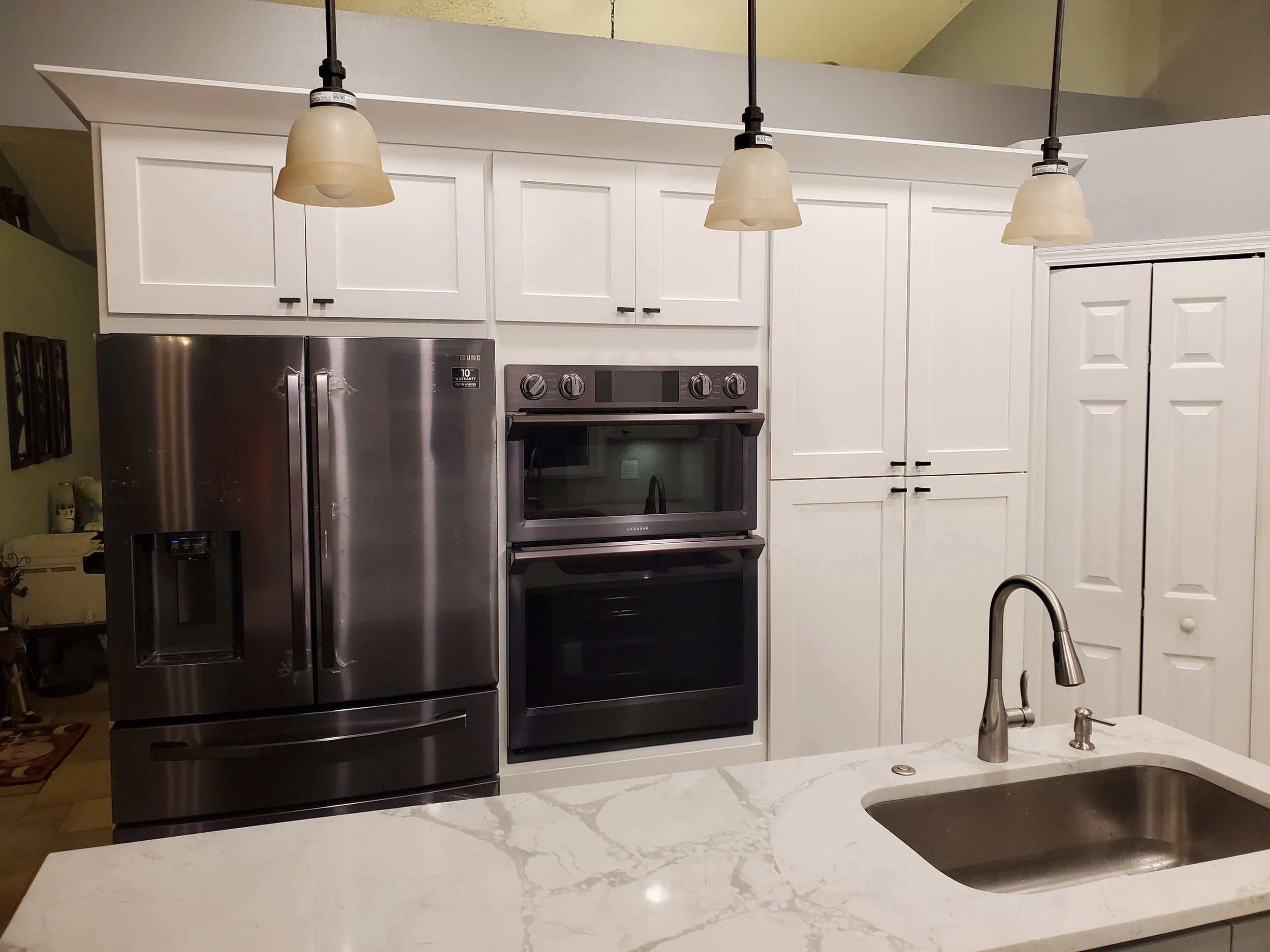 Modern kitchen with stainless steel refrigerator, double oven, white cabinets, and a marble countertop with a sink.