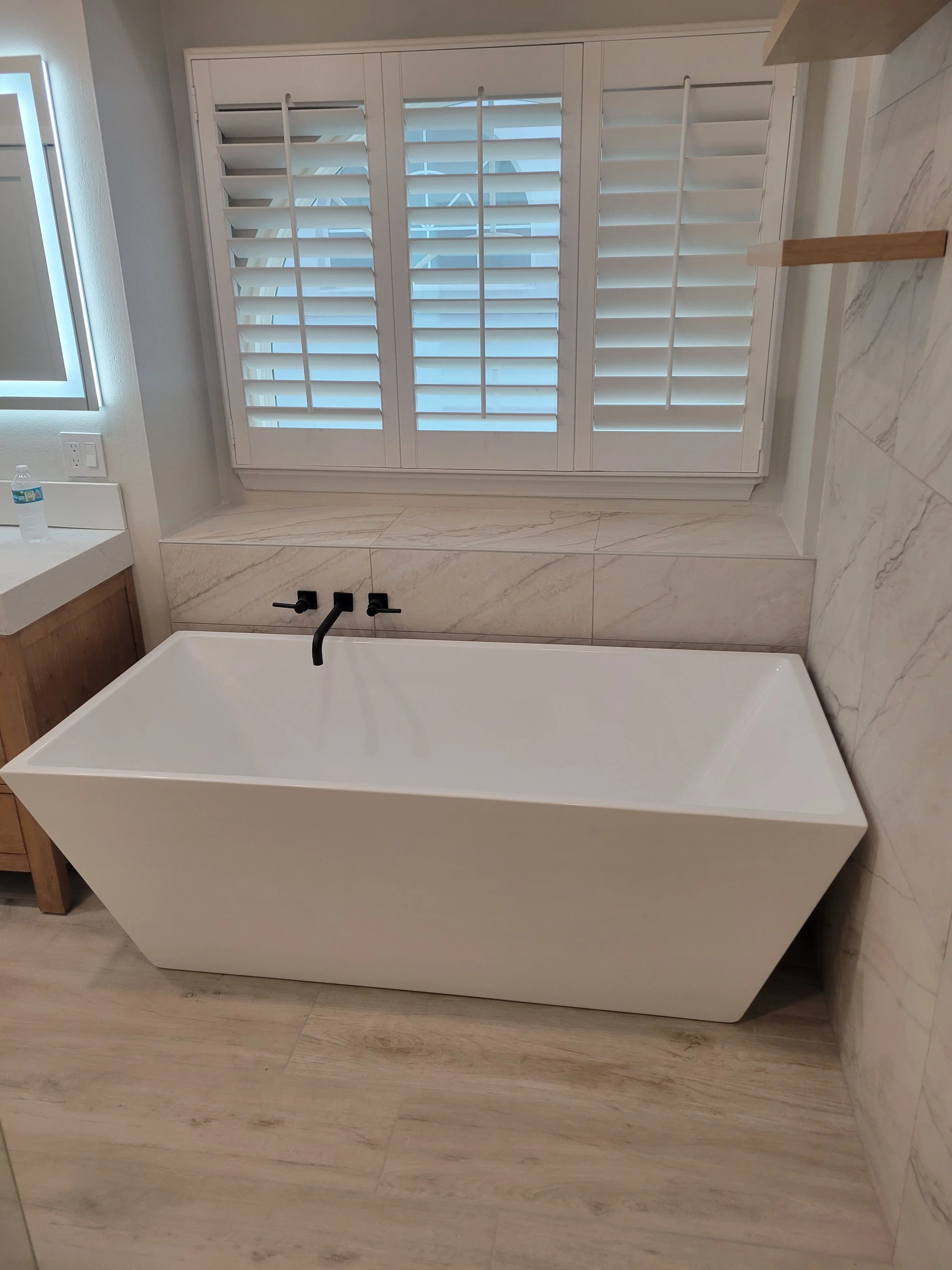 White modern bathtub with black fixtures in a bathroom with light beige wood-look tile flooring and wall tiles, and white plantation shutters above.
