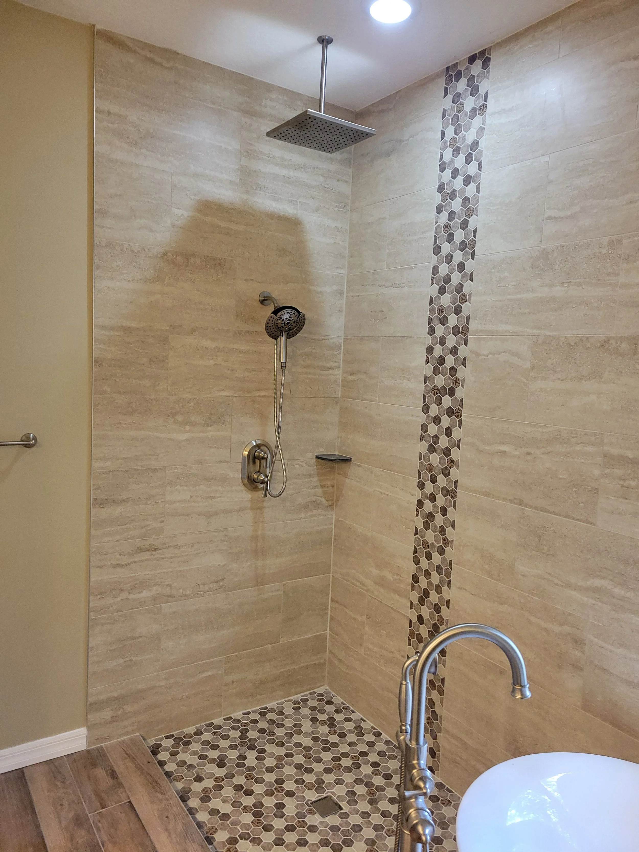 A bathroom shower area with beige and brown tile walls, a rainfall showerhead, a handheld showerhead, a small shelf, and a tiled floor. Part of a bathtub with a silver faucet is visible in the foreground.