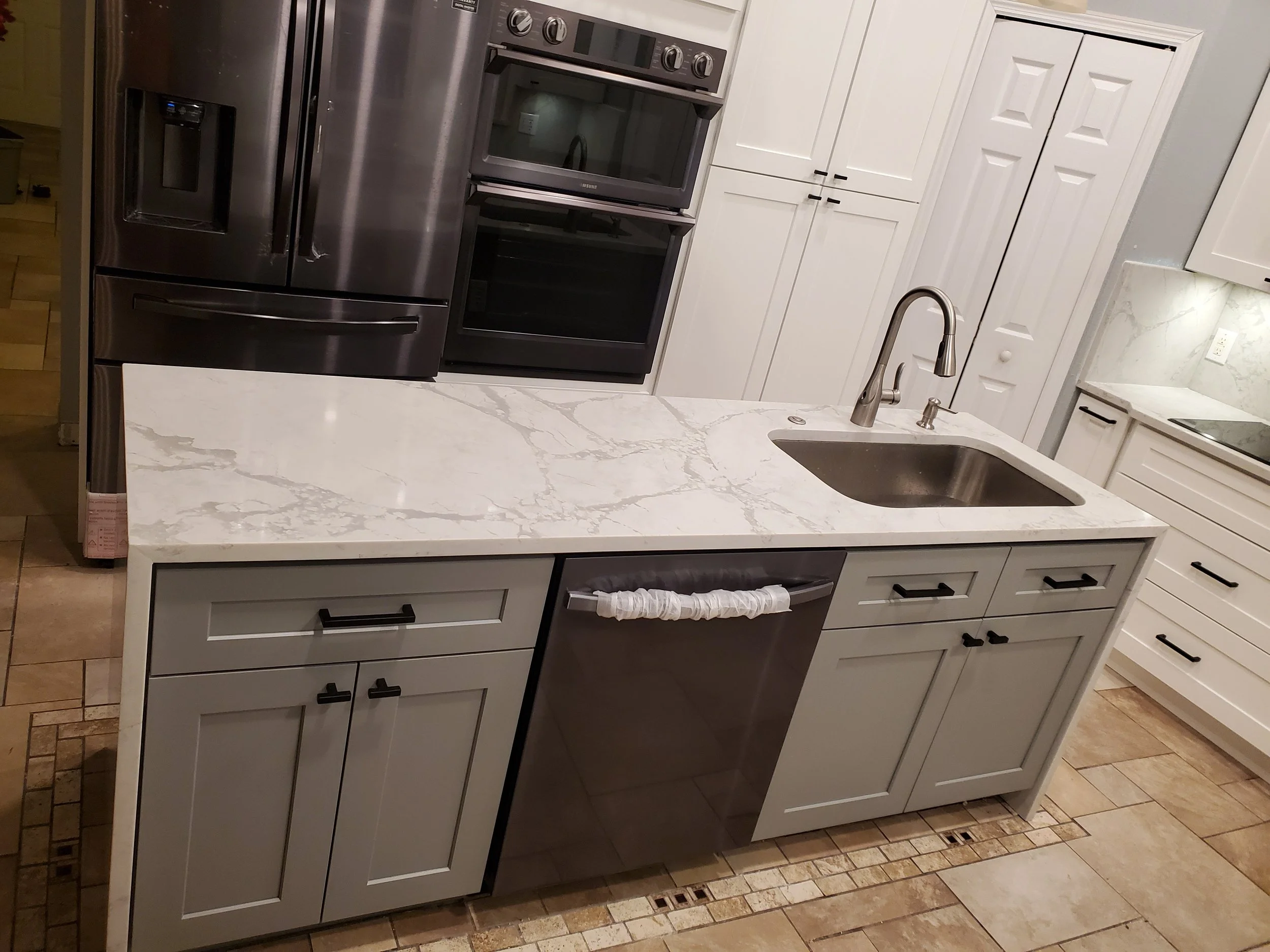 Modern kitchen with marble countertop island, stainless steel refrigerator, built-in oven, gray cabinets with black handles, and a sink with a tall faucet.