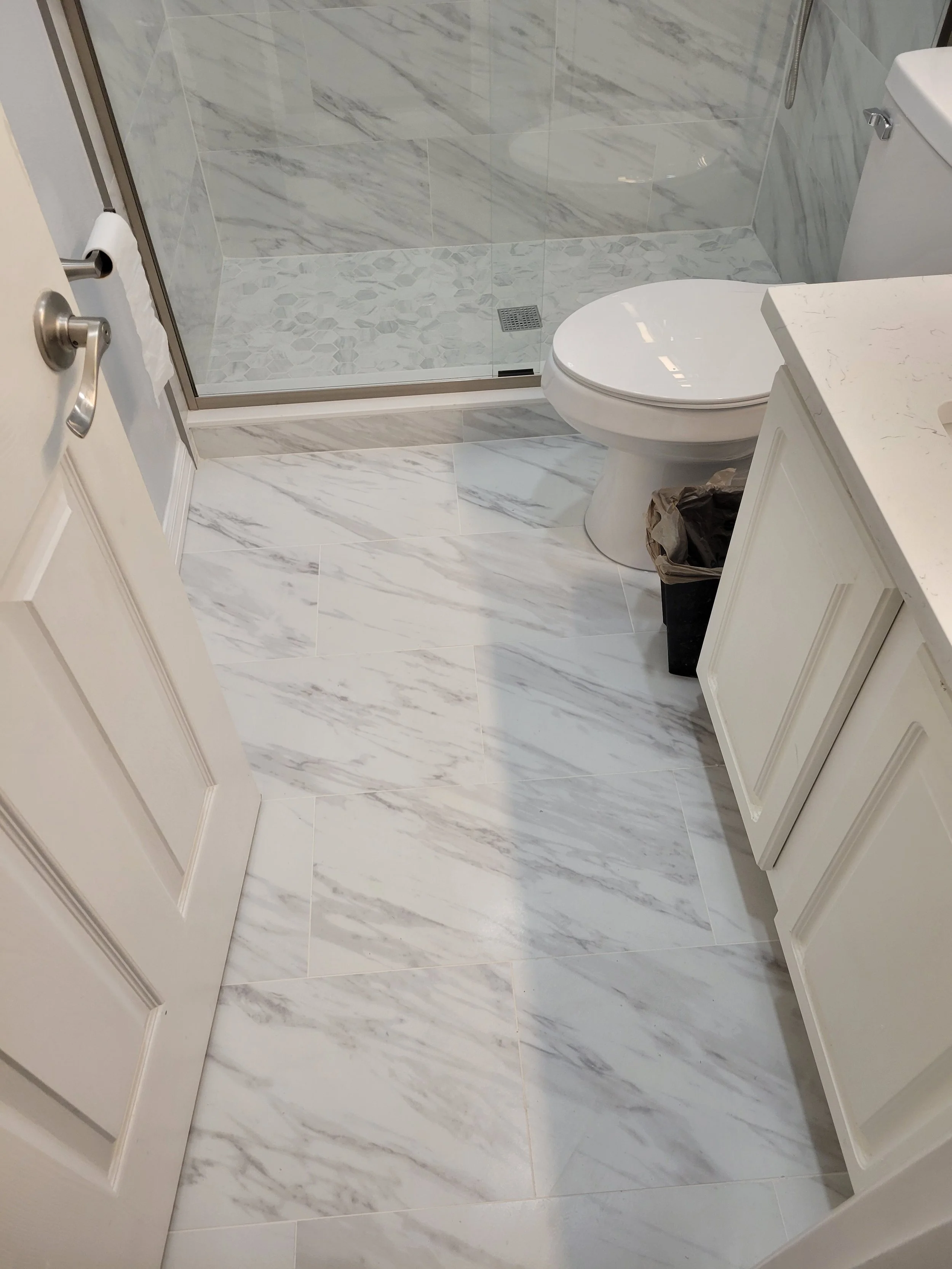 The bathroom has marble tile flooring and walls, a glass-enclosed shower with a drain, and a white vanity with a marble countertop