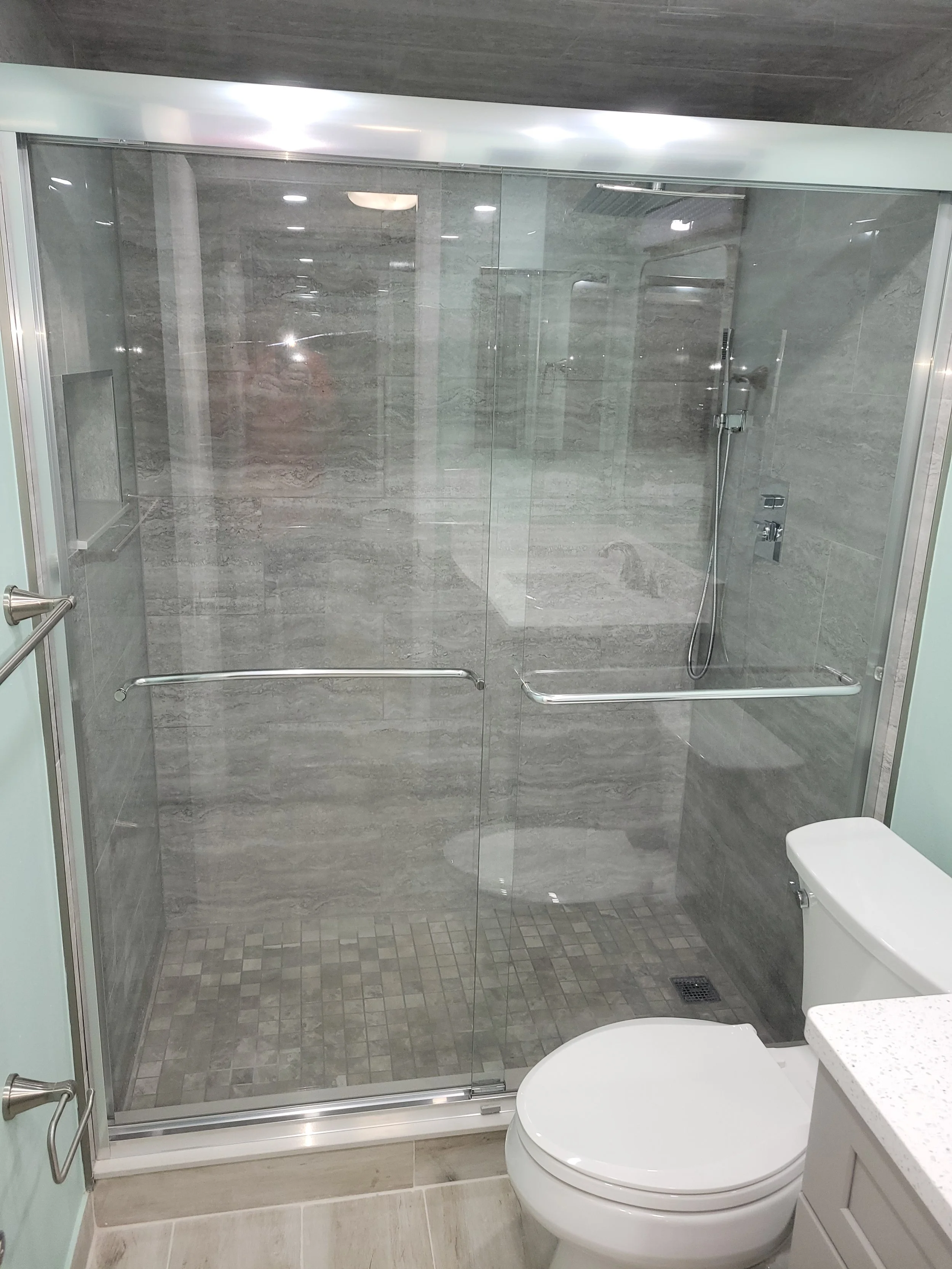 A modern bathroom with a glass-enclosed shower featuring gray marble tiles, a hand-held showerhead, and a built-in shelf, next to a white toilet and a light-colored vanity with a granite countertop.
