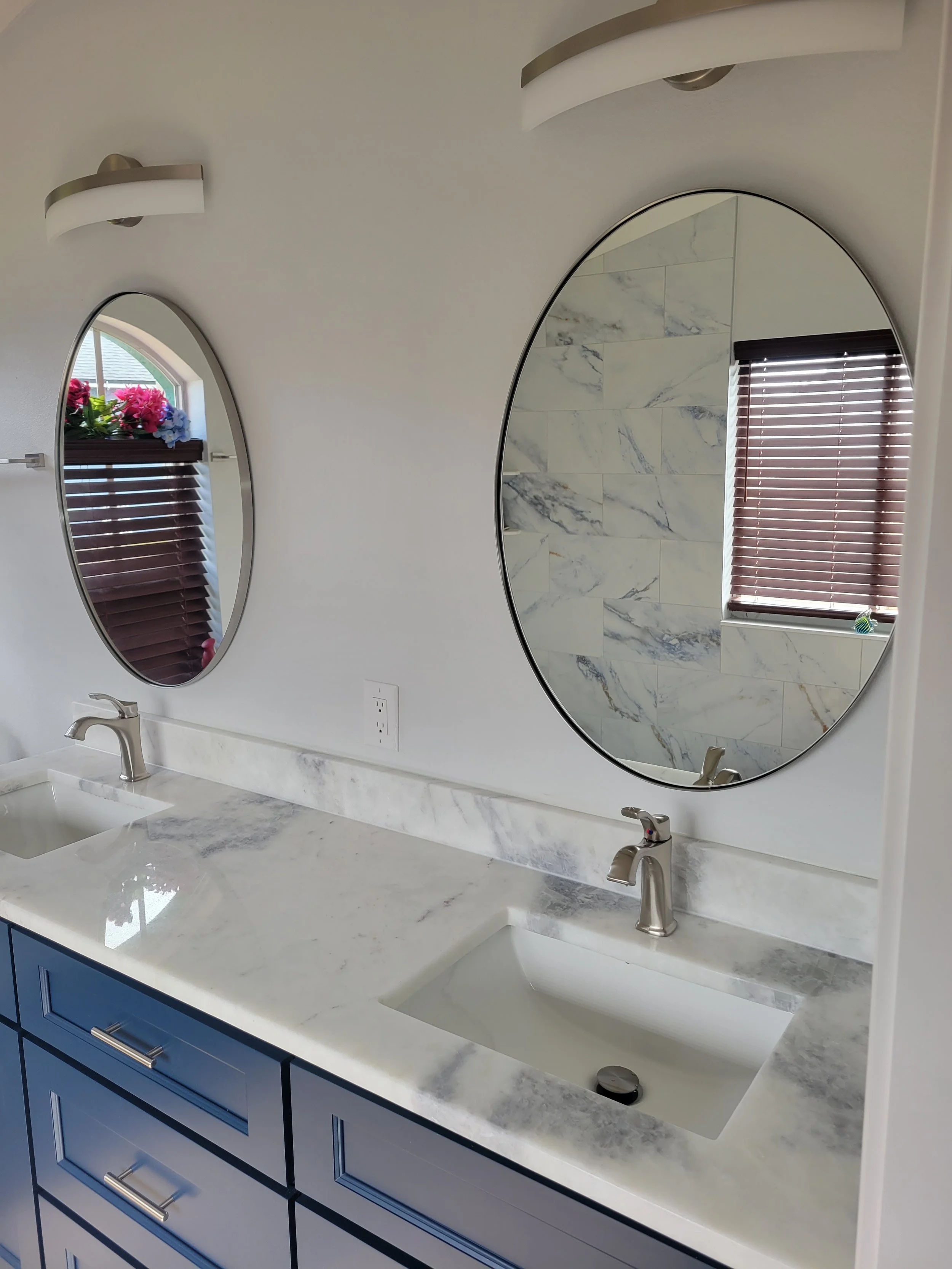 Bathroom vanity with two oval mirrors, marble countertop, and two sinks with silver faucets. There are wooden blinds in the window and a small potted plant on the windowsill.