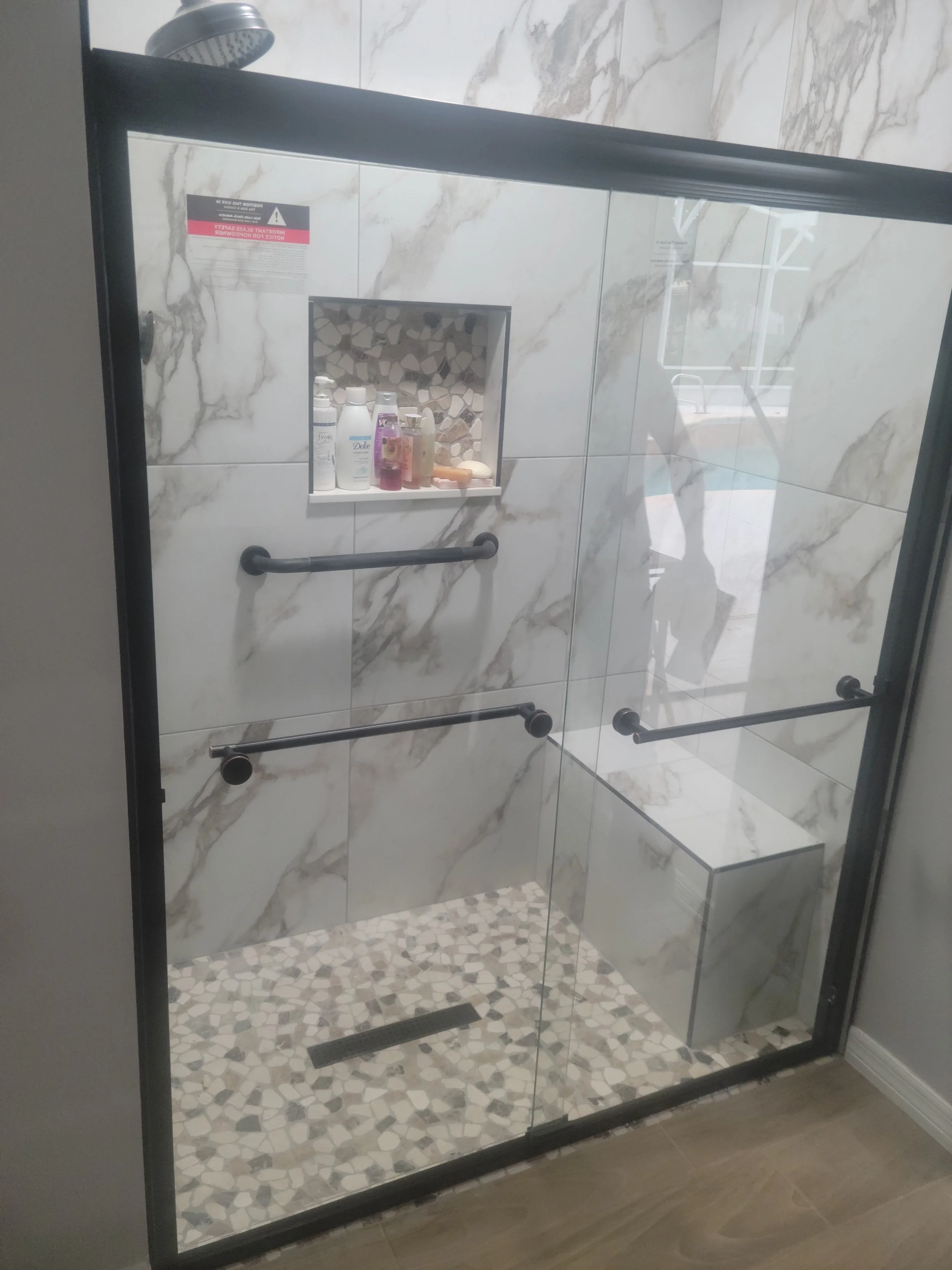 A modern, glass-enclosed shower with white marble walls, a built-in niche with toiletries, a bench, and black grab bars.