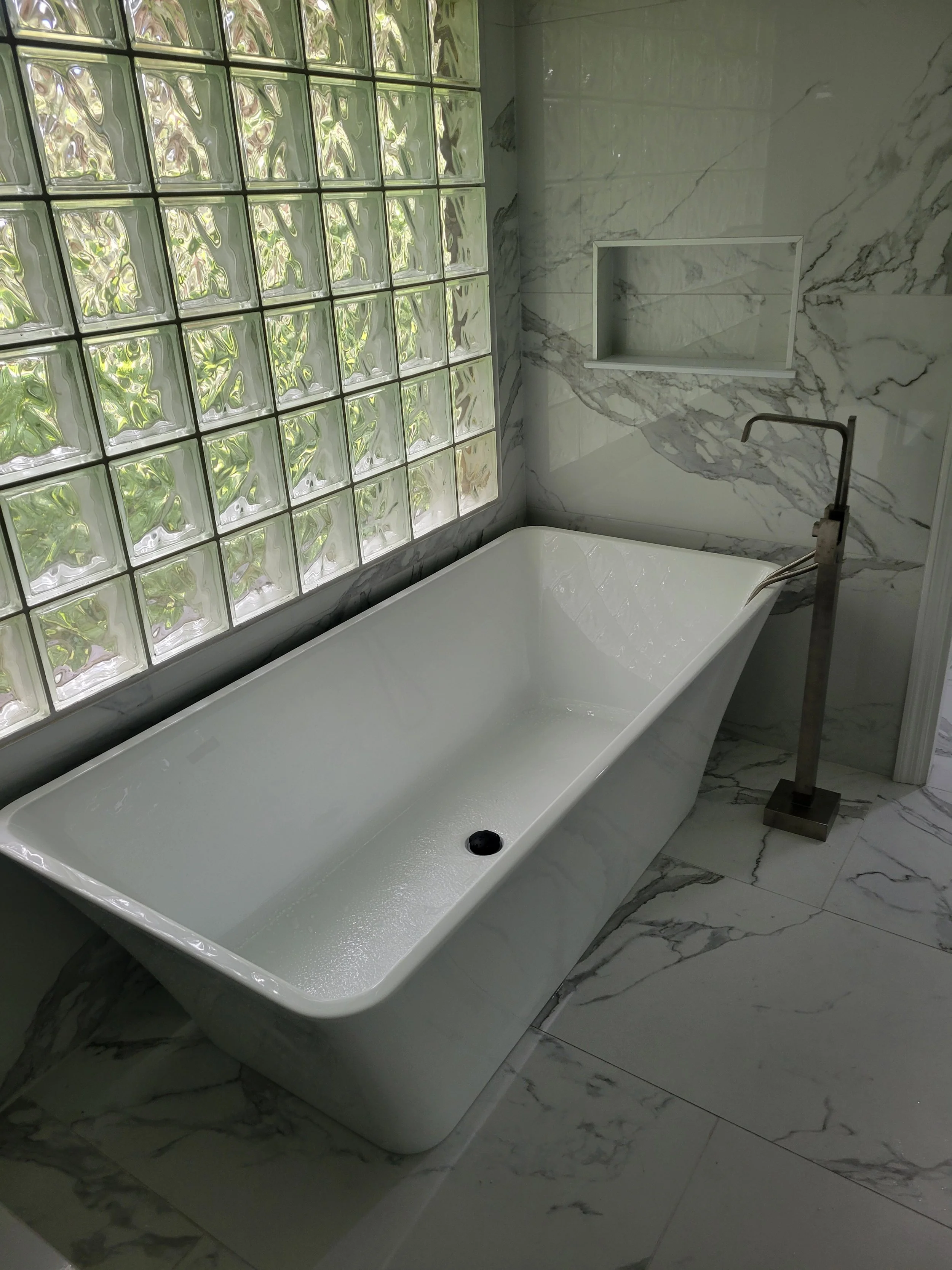 Modern bathroom with a white freestanding bathtub, marble walls, a glass block window, and a tall bathtub faucet.
