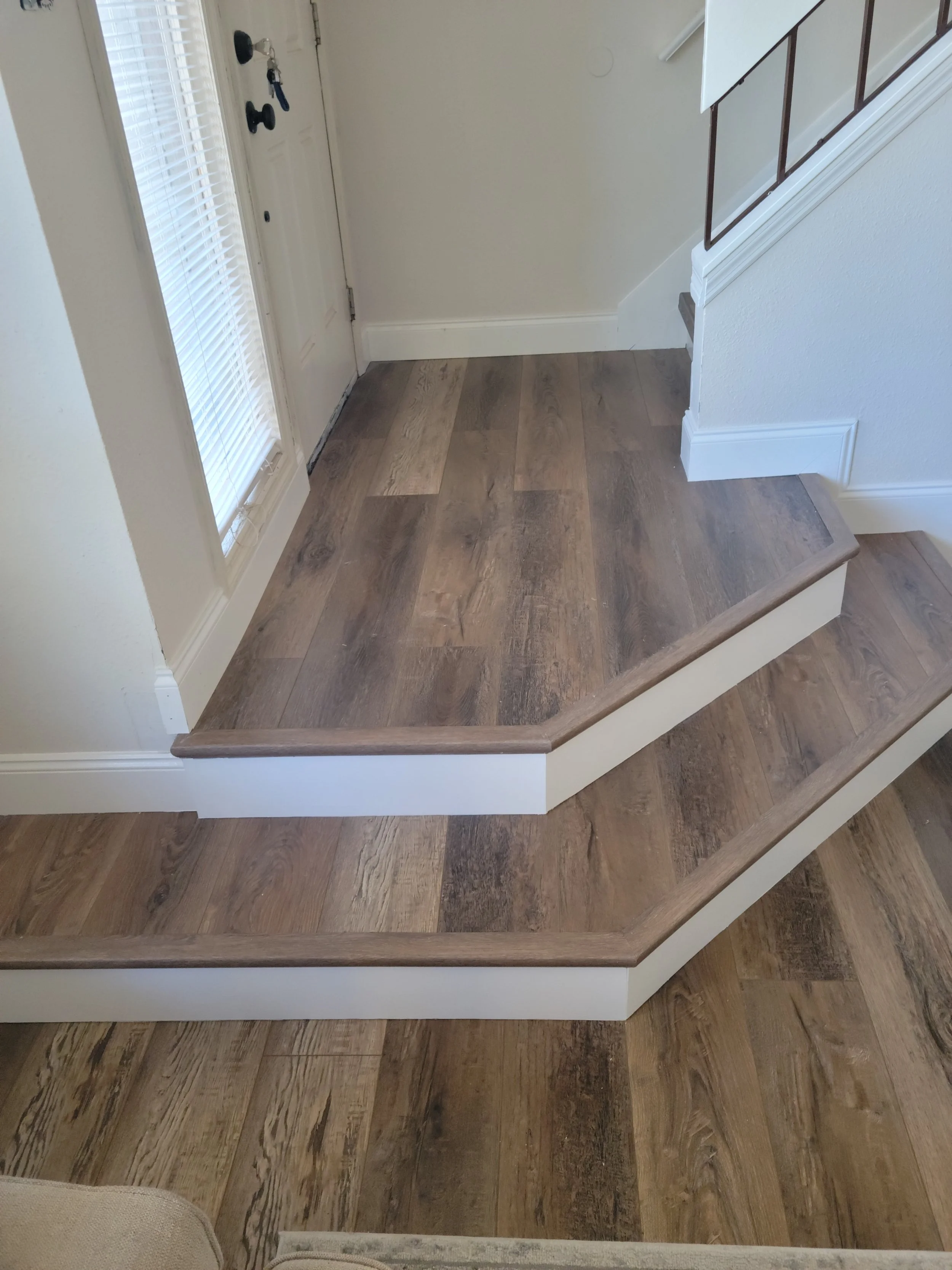 Foot of stairs with a landing, hardwood flooring, and white baseboards near an entry door with blinds.