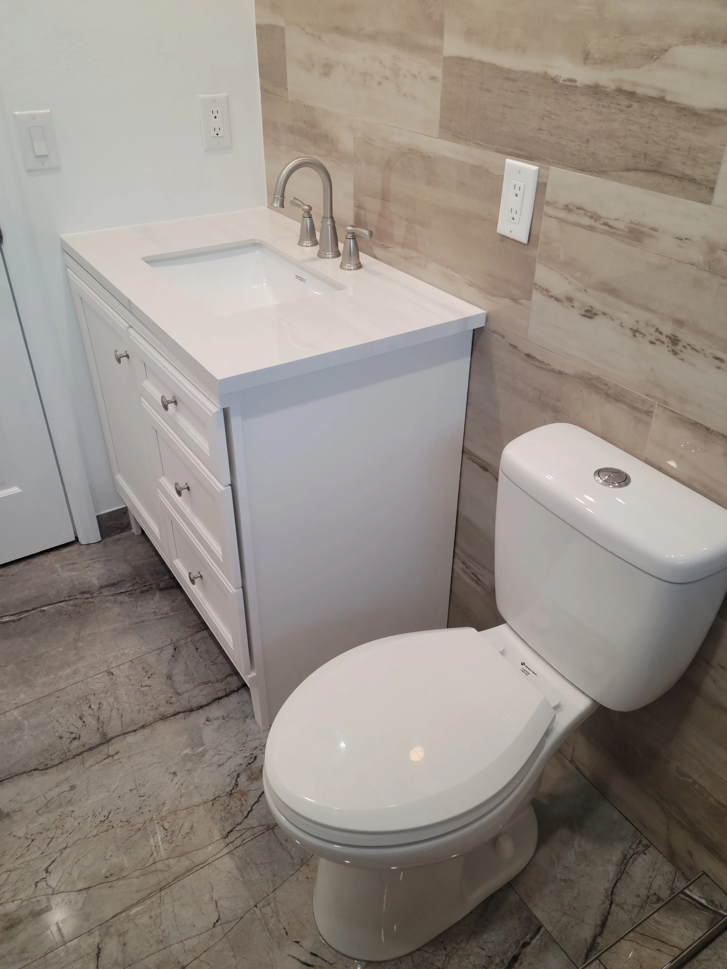 Bathroom with a white vanity sink, a silver faucet and handles, a white toilet, and beige/stoney wall tiles.