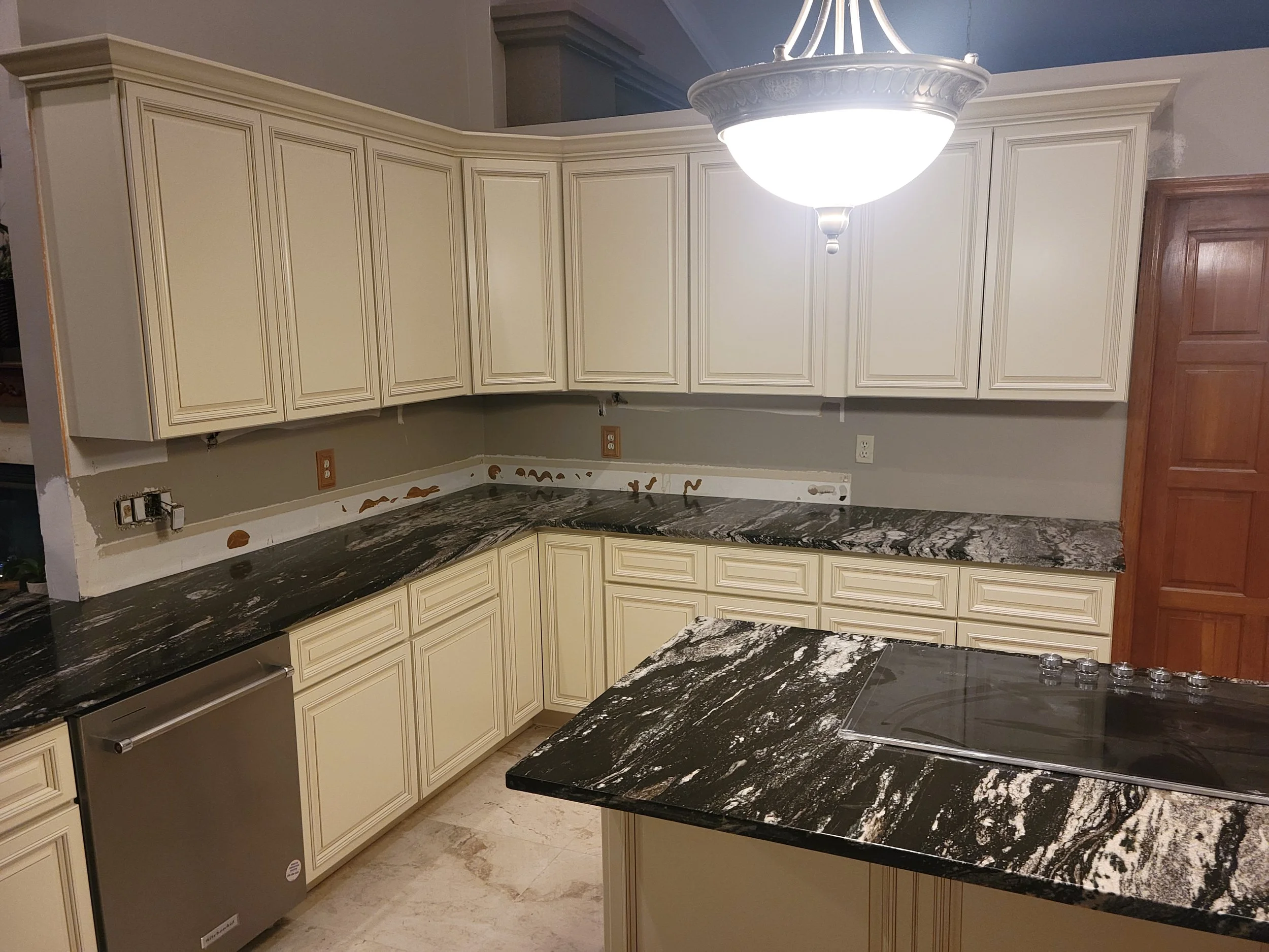 Kitchen with cream-colored cabinets, black and white marble countertops, stainless steel dishwasher, unfinished wall area, and a wooden door.