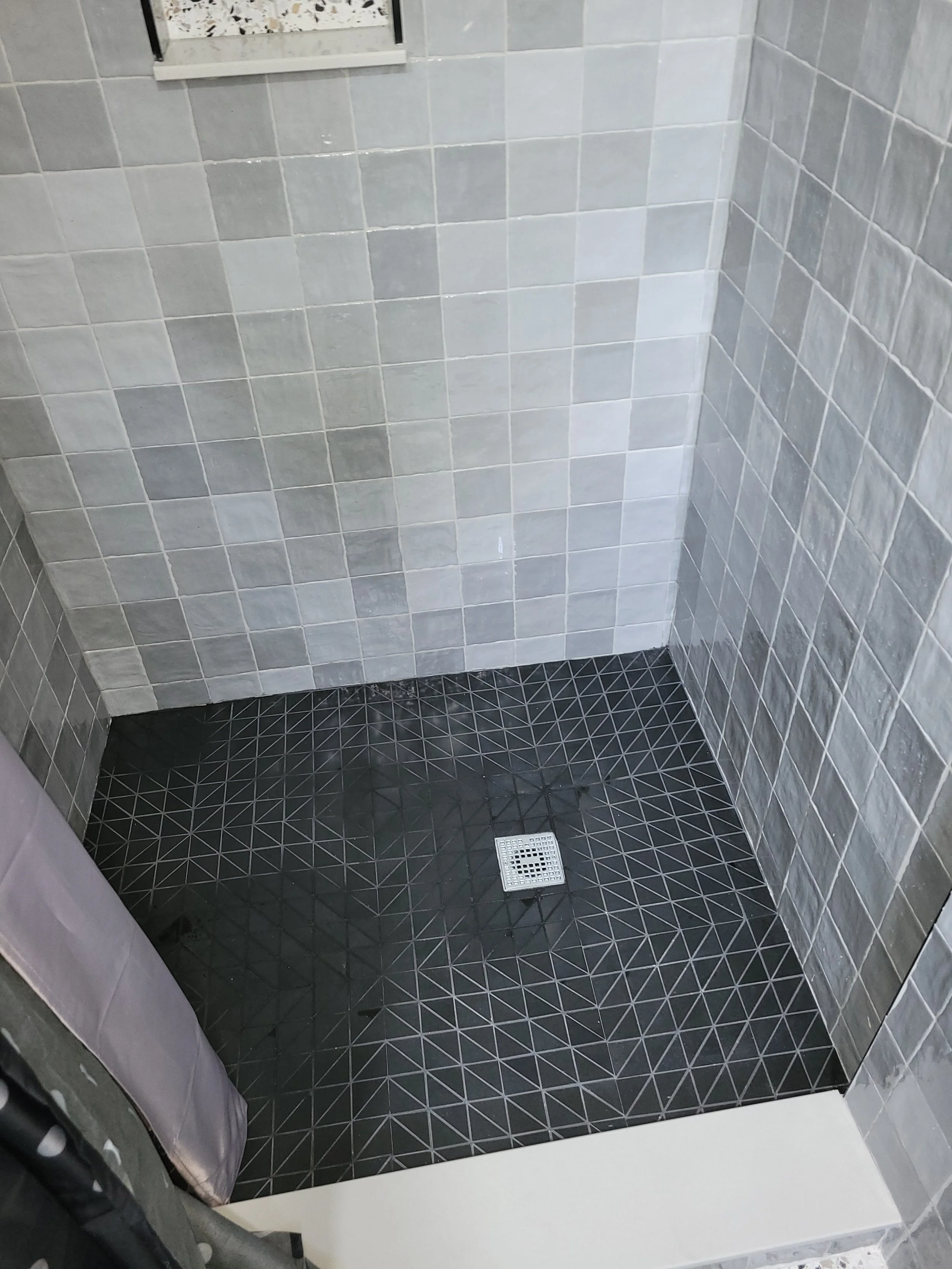 Shower area with black geometric floor tiles, grey wall tiles, and a drain in the center of the floor.