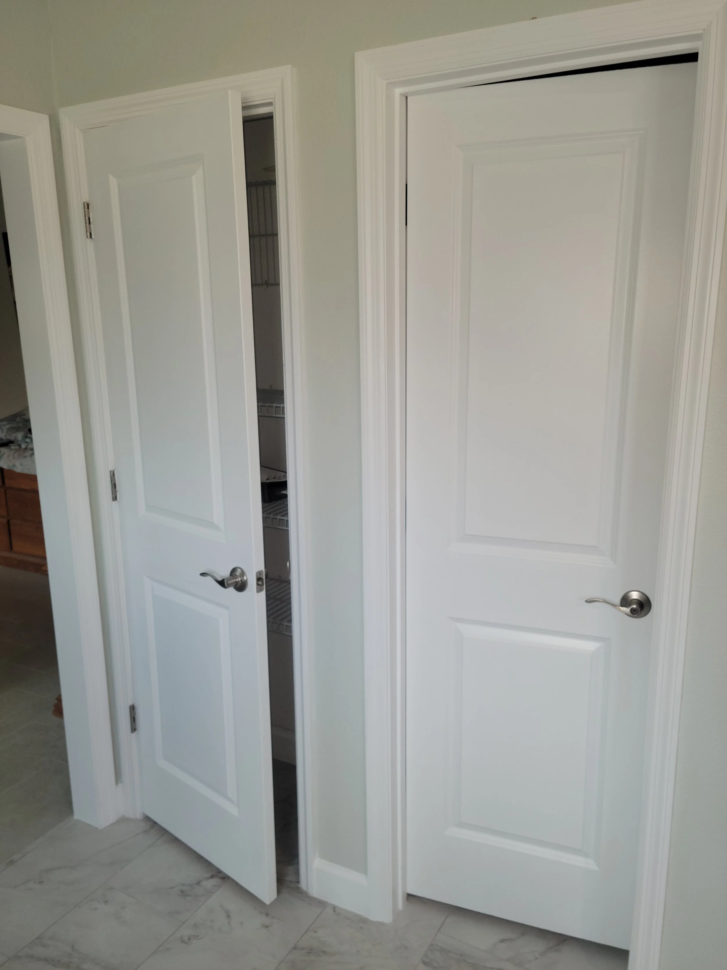 Interior view of two white doors, one partially open revealing a closet with wire shelves, in a room with light-colored walls and marble flooring.