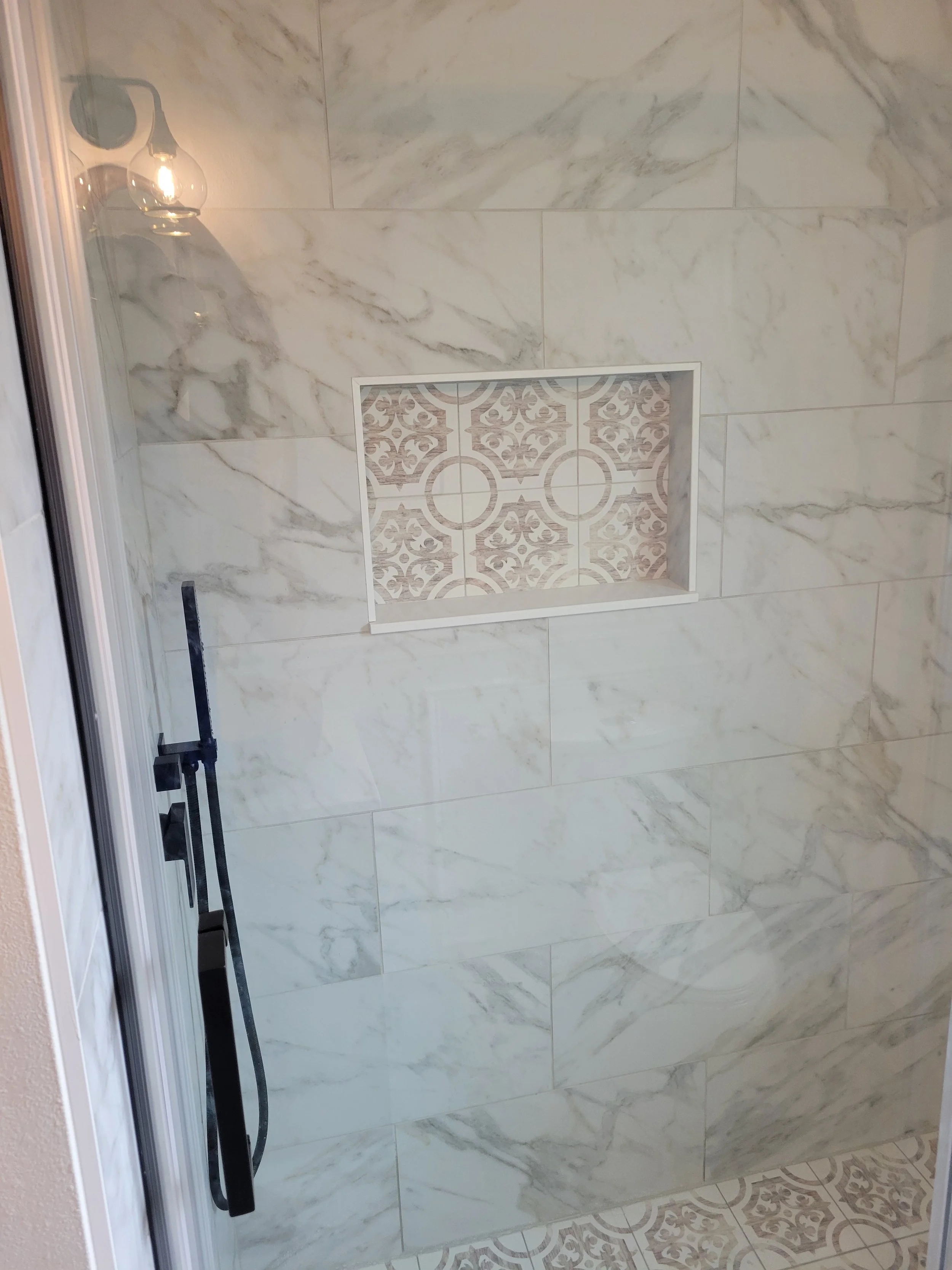 A marble tiled shower with an inset decorative tiled niche and a black shower handle.