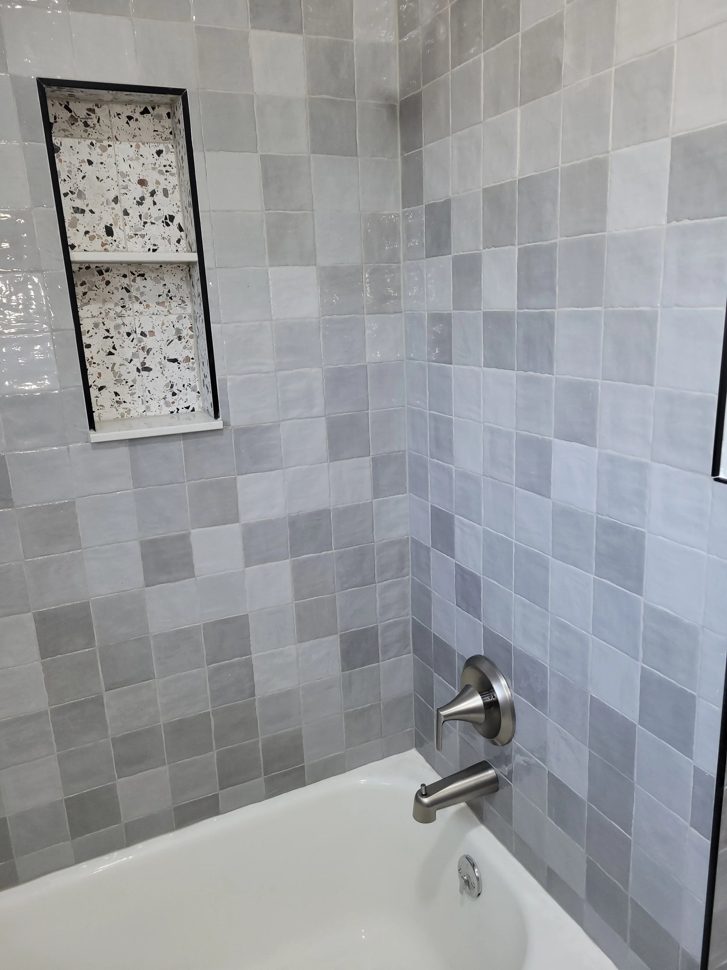 Bathroom corner with tiled wall, built-in shelf, and modern silver faucet and handle on a bathtub.
