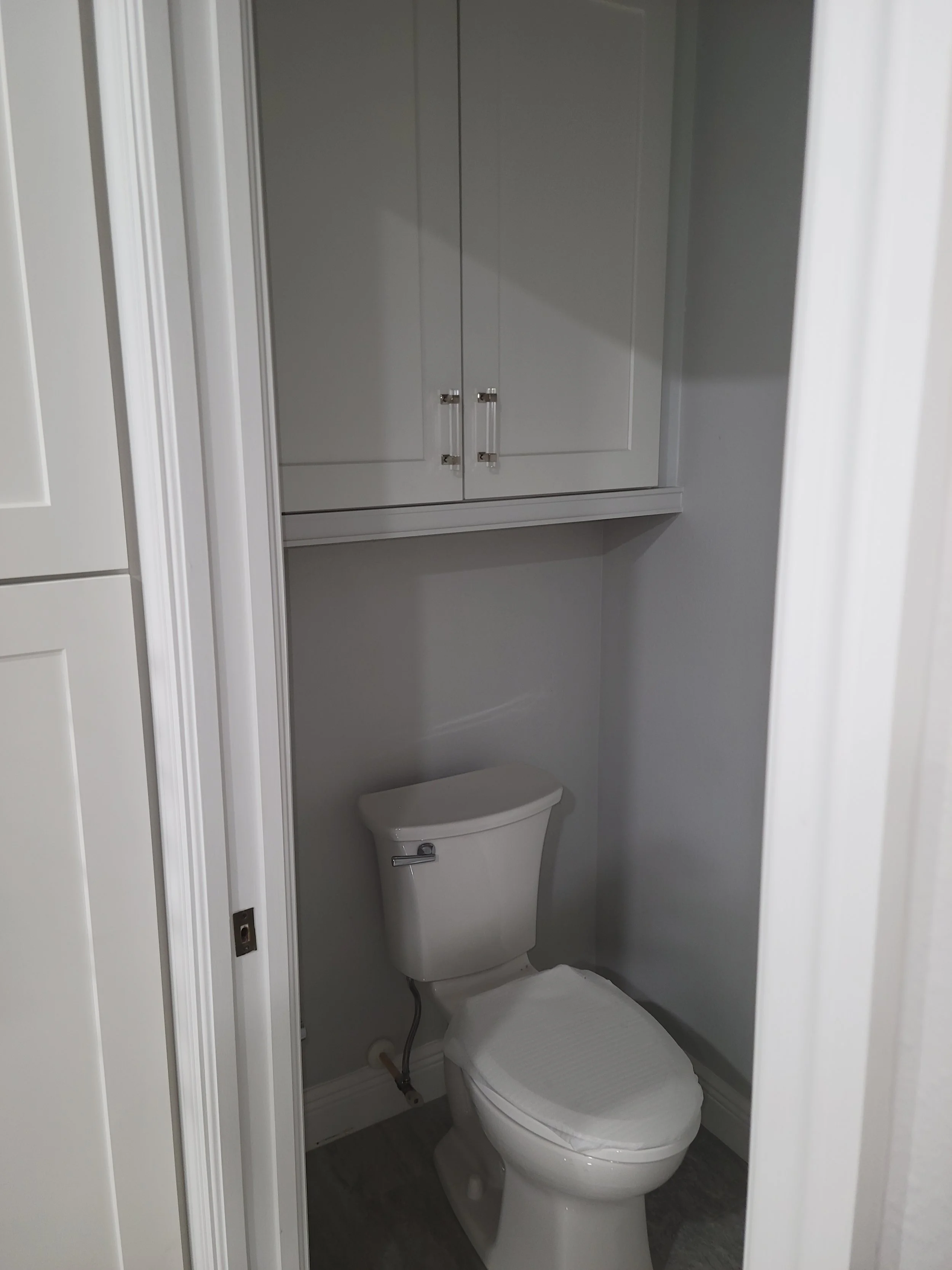 Small bathroom with a white toilet and a white cabinet above it, gray walls, gray flooring, and white trim.