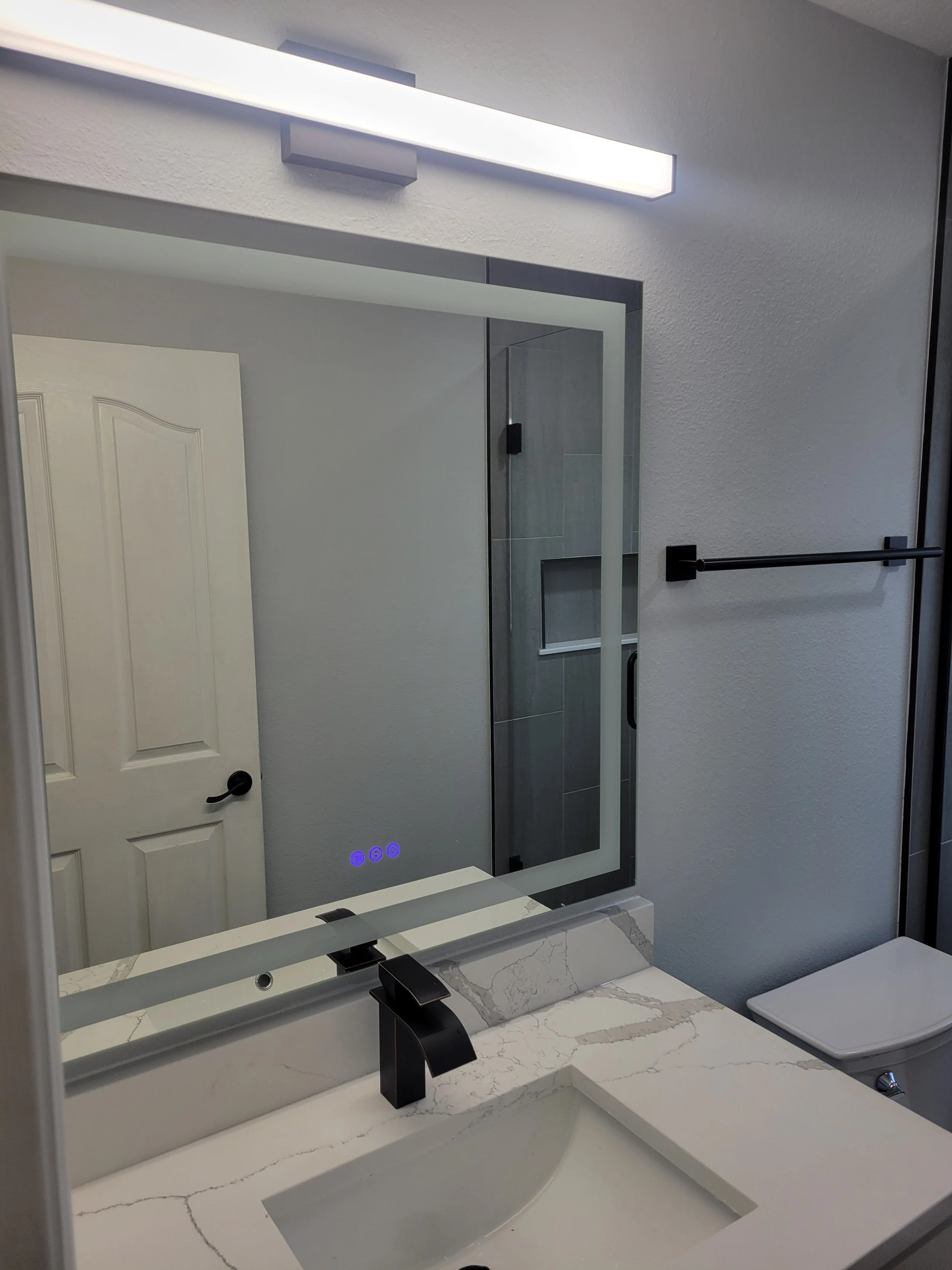 Modern bathroom vanity with marble countertop, black faucet, large mirror, and a light fixture above the mirror. Part of a shower with tiled walls and a glass door is visible in the reflection.