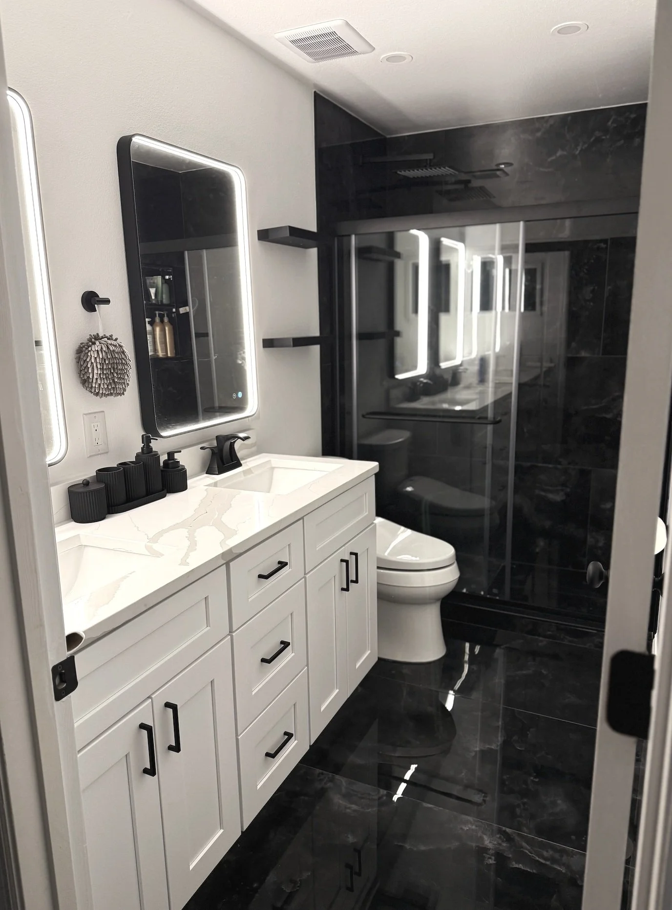 After image of a modern bathroom equipped with black hardware, illuminated vanity mirrors, black marble floors, and a glass shower enclosure with black marble walls.