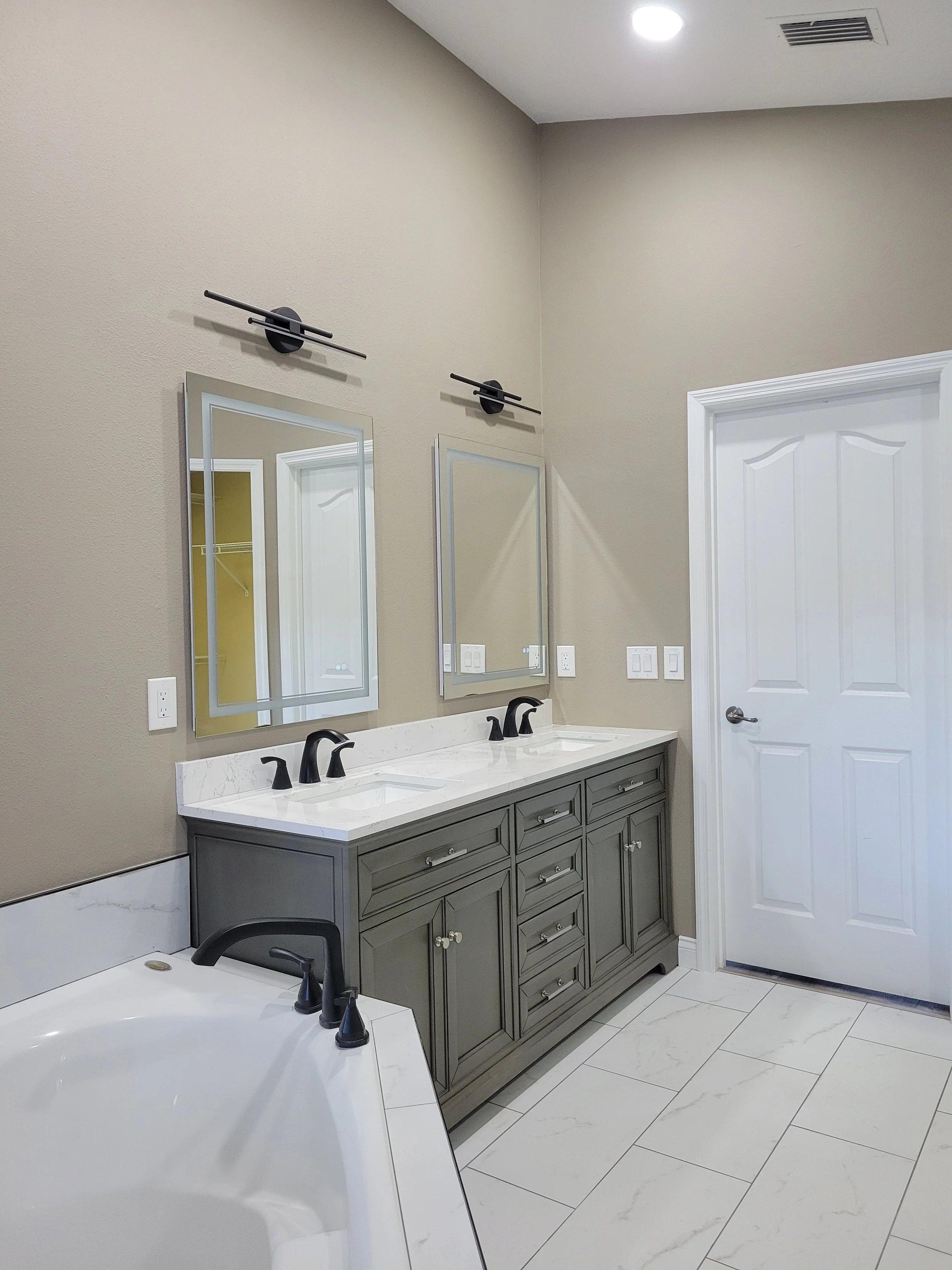 Bathroom with double vanity, gray cabinets, white marble countertops, two mirrors, black fixtures, white tile flooring, next to a bathtub, and a closed white door.