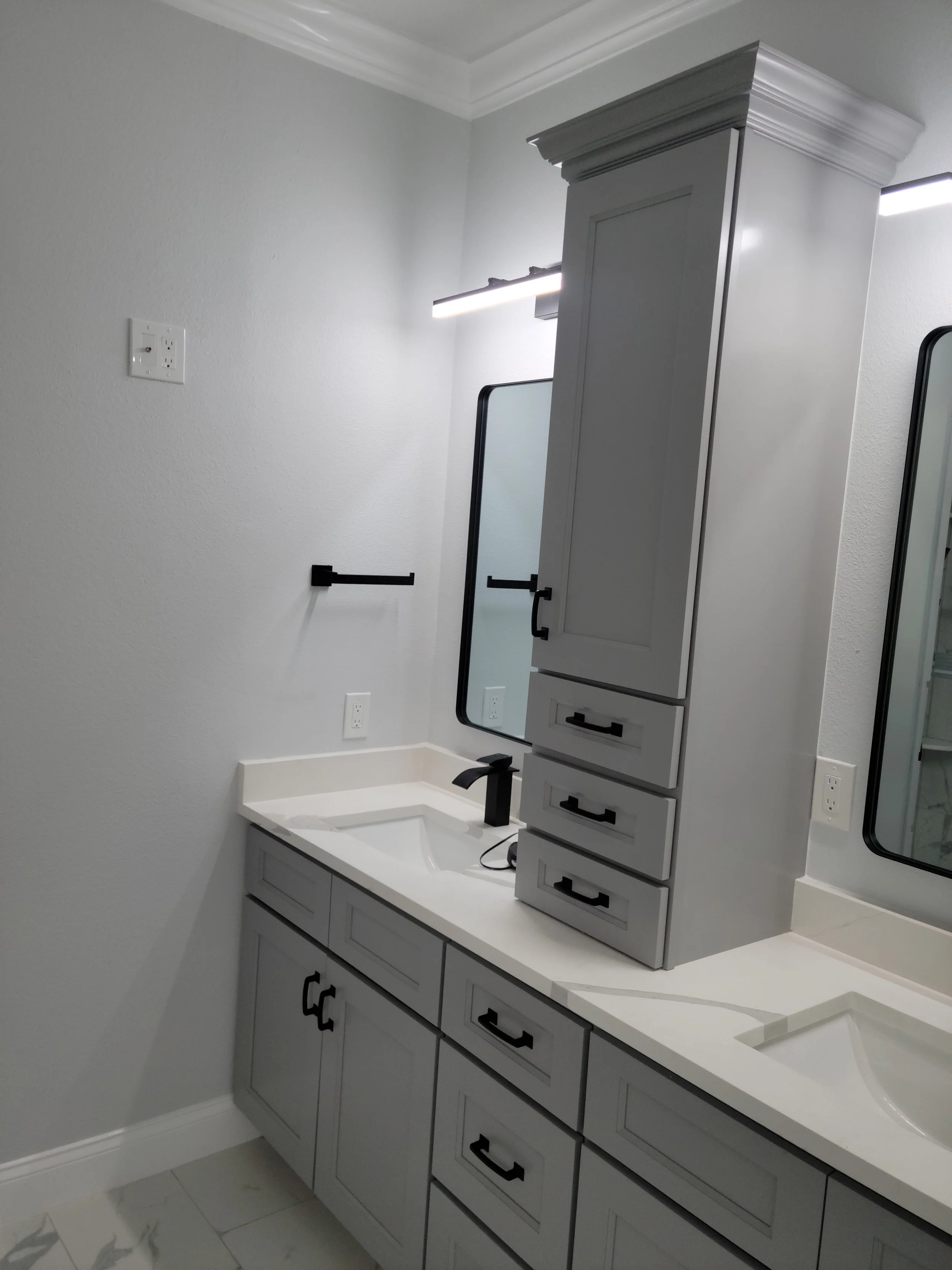 Gray bathroom vanity with two sinks, black fixtures, a tall cabinet, and two mirrors with black frames on a white wall.