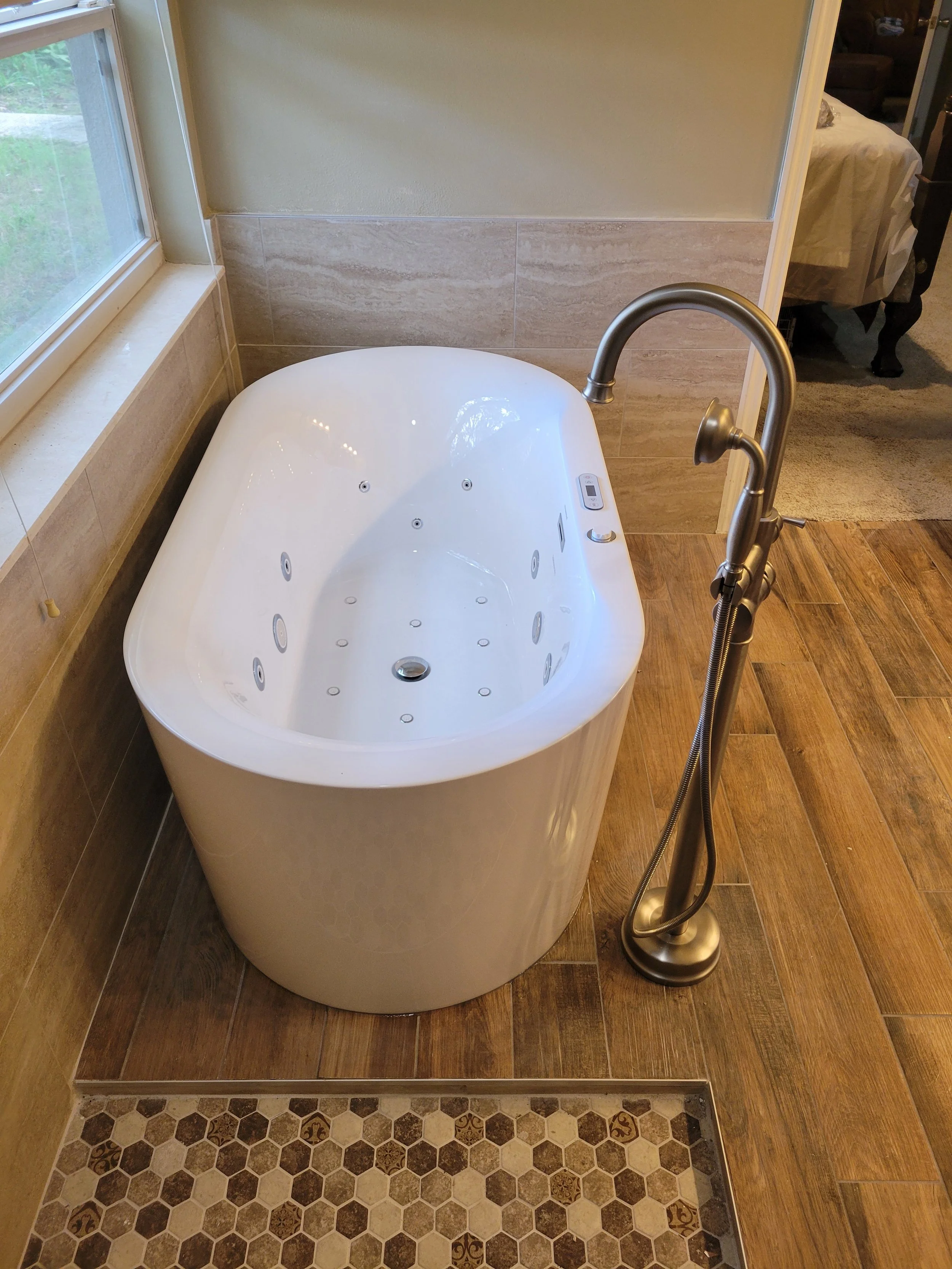 A modern white freestanding bathtub with multiple jets, positioned near a window, with a tall silver faucet and handheld showerhead, on a wooden-floored bathroom with beige and brown tiles.