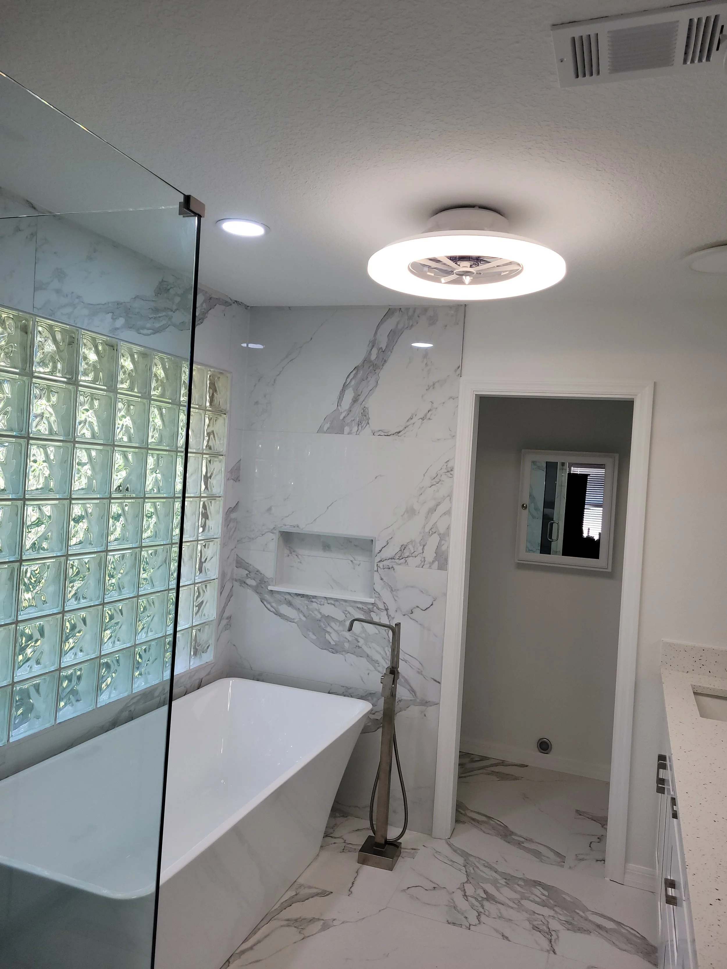 Modern bathroom with white marble walls and floor, standalone bathtub, glass block wall, and ceiling light fixture.