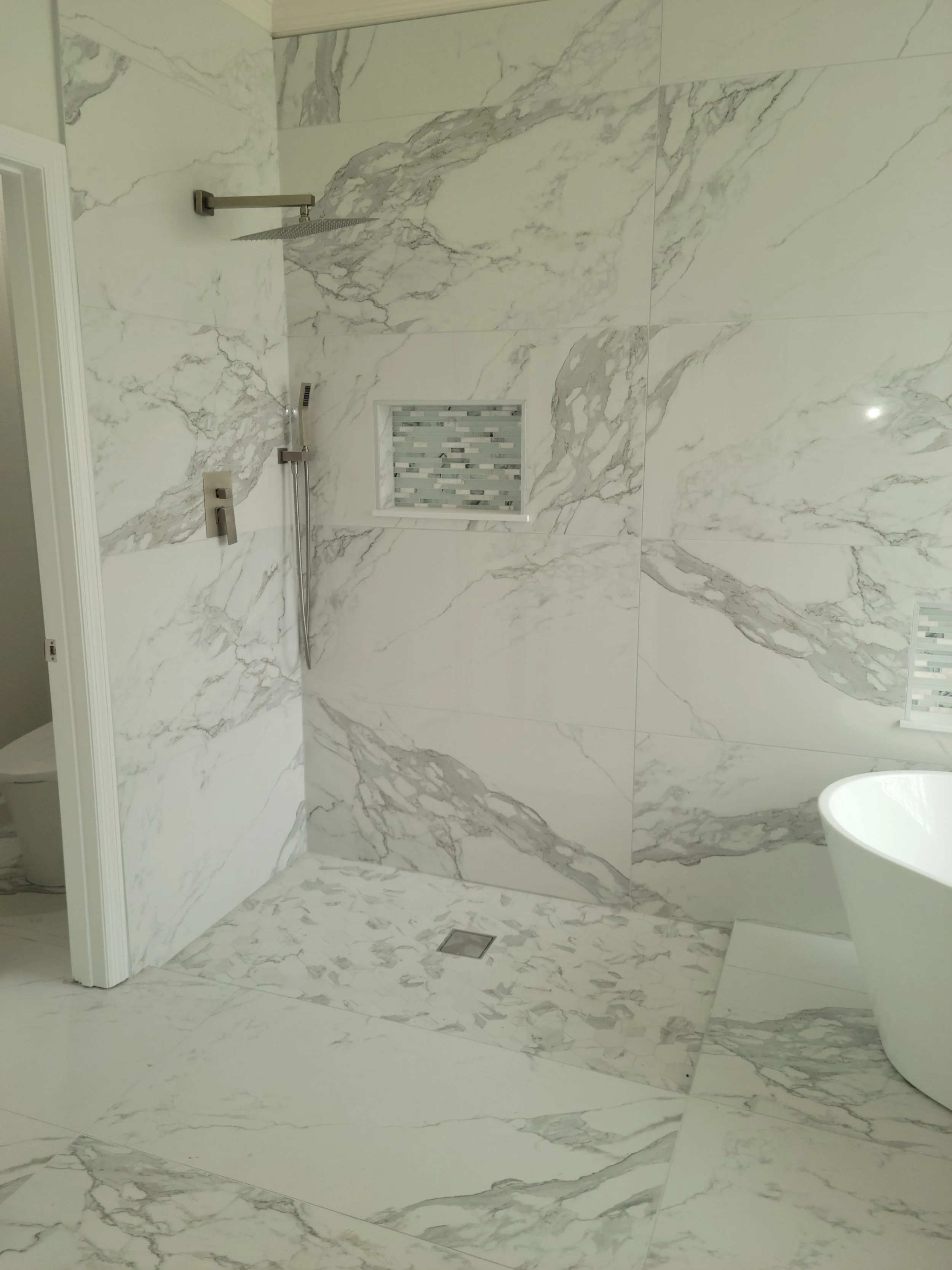 Bathroom shower area with marble tiles, built-in shower niche, rainfall showerhead, handheld shower, and a white freestanding bathtub.