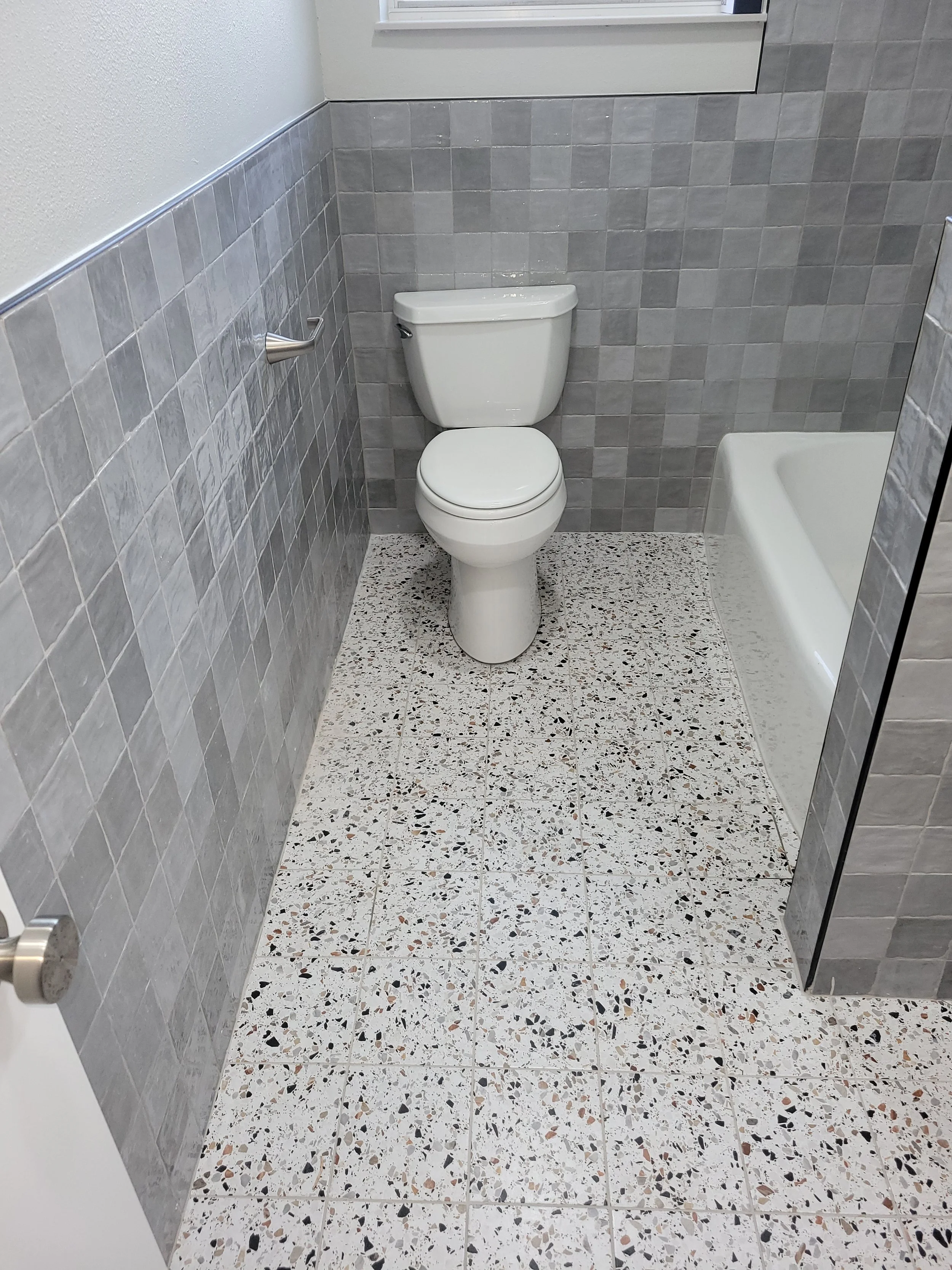 Bathroom with a white toilet, grey tiled wall, porcelain floor tiles, and a partial view of a bathtub.
