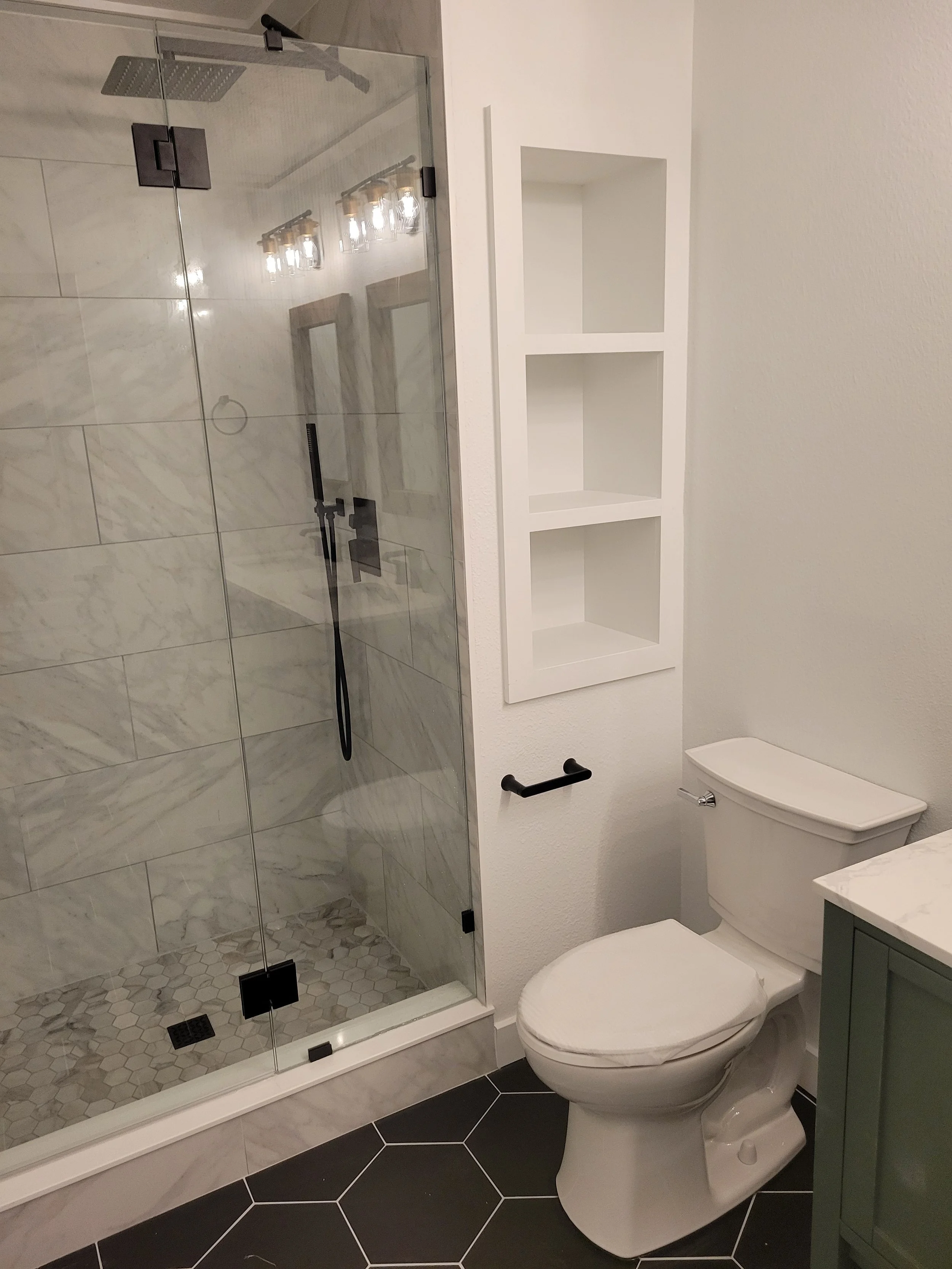 Bathroom with a glass shower enclosure, white shelving unit, and toilet with a closed lid, black hexagonal floor tiles.