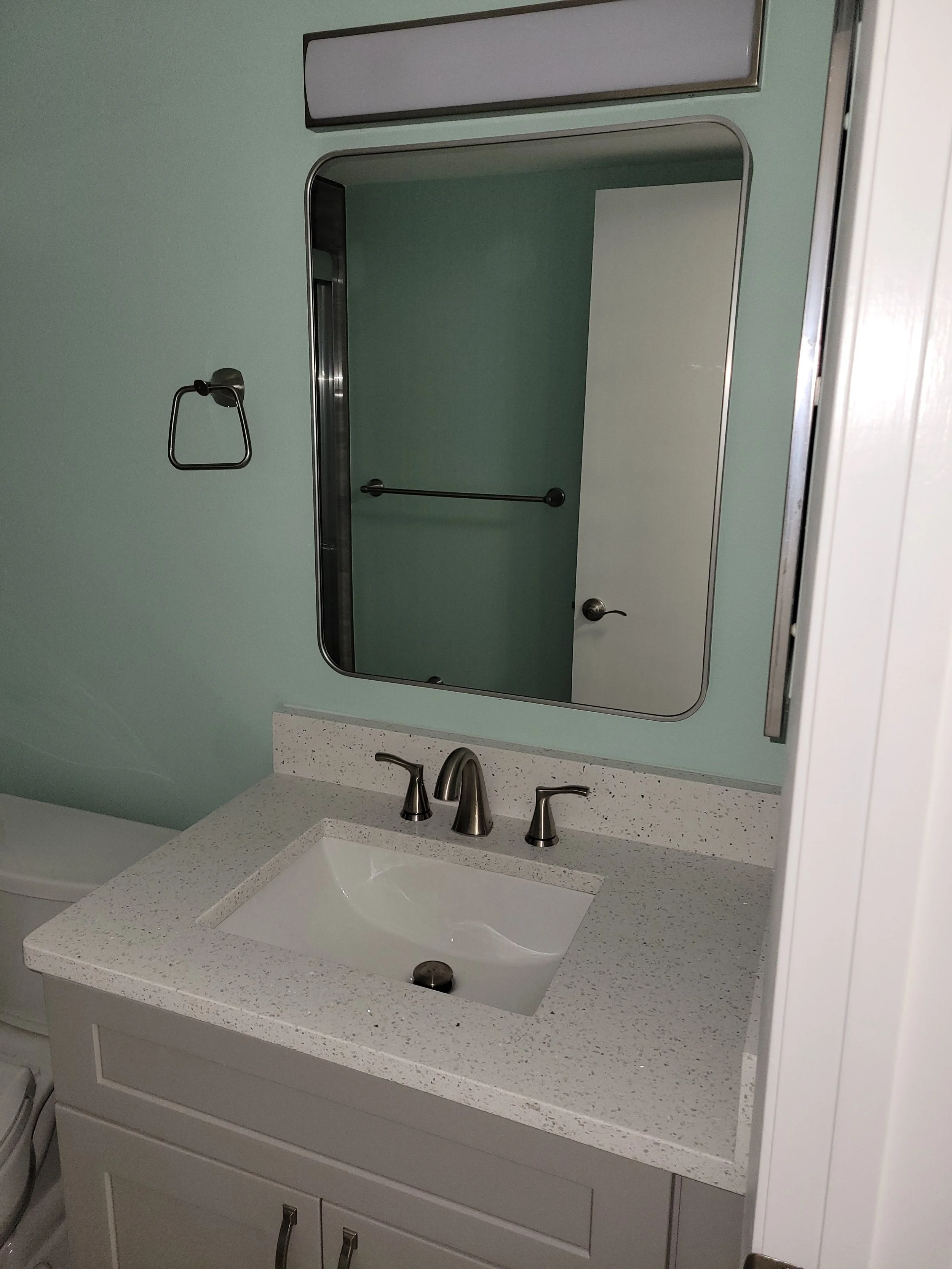Bathroom sink with a granite countertop and a rectangular mirror above it, light green wall, and part of a white cabinet below.