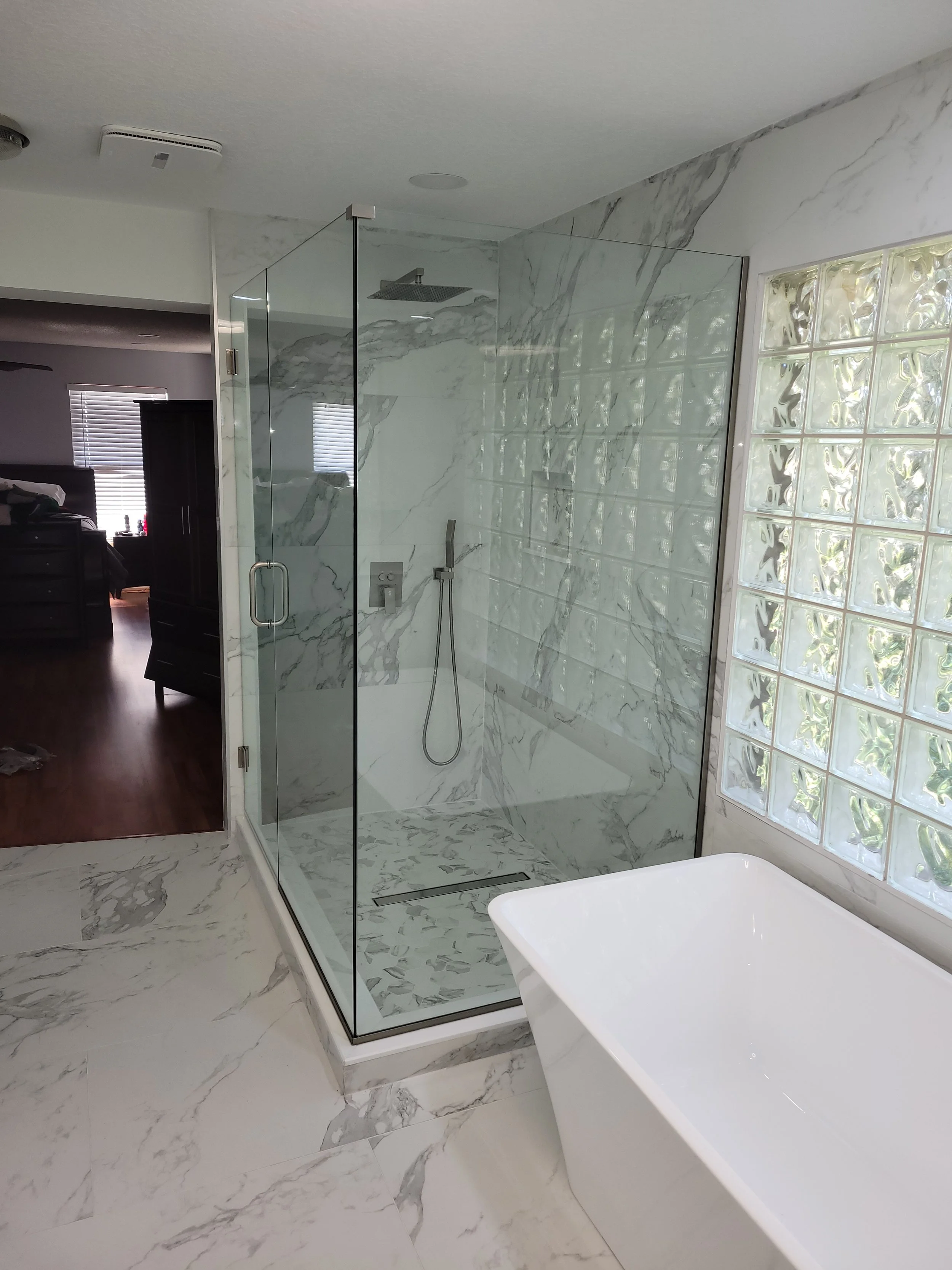 Modern bathroom with marble floors, a glass-enclosed shower with marble walls, a white bathtub, and a glass block wall allowing natural light.