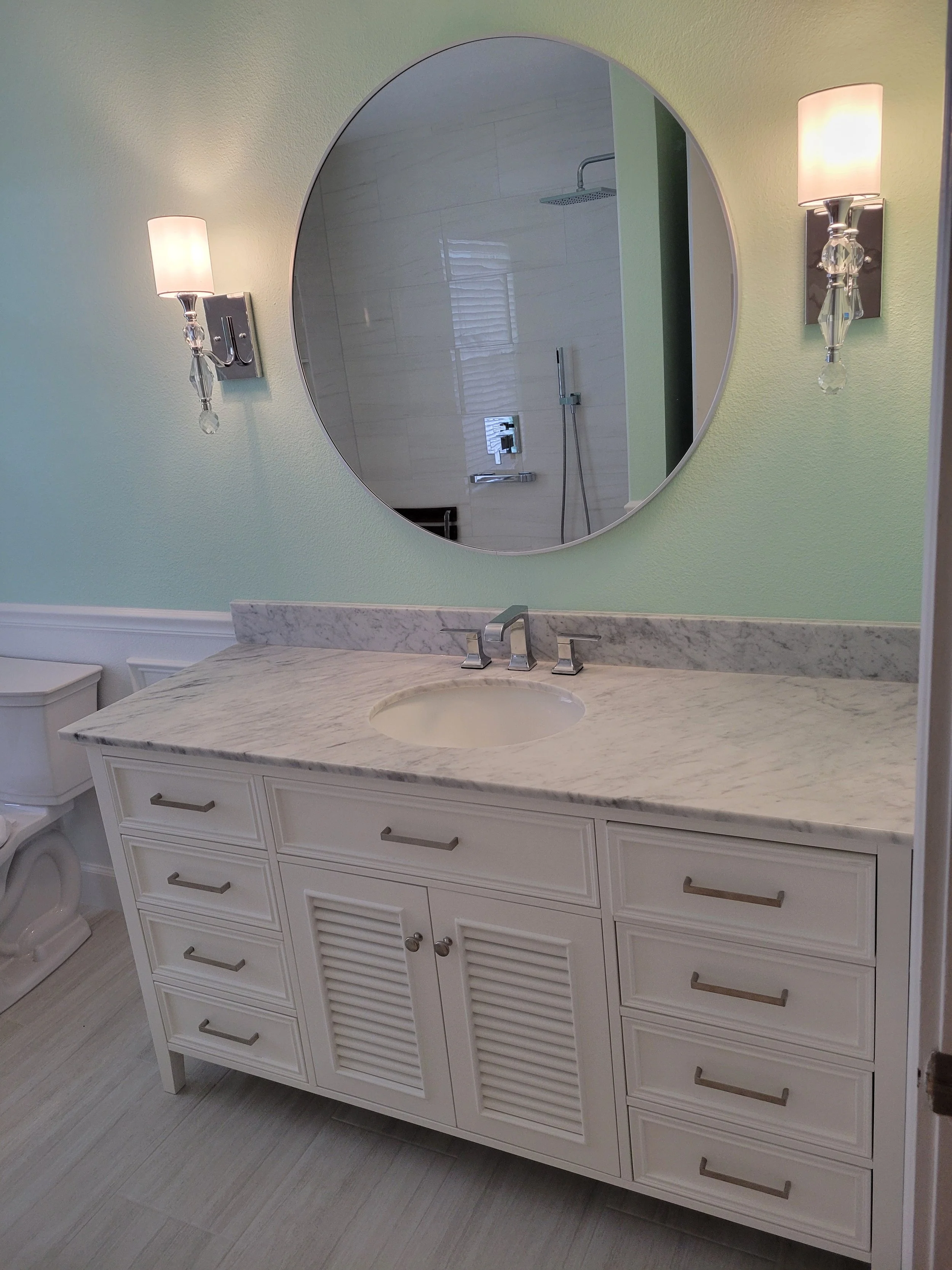 Bathroom vanity with marble countertop, white cabinetry, round mirror, and wall-mounted light fixtures