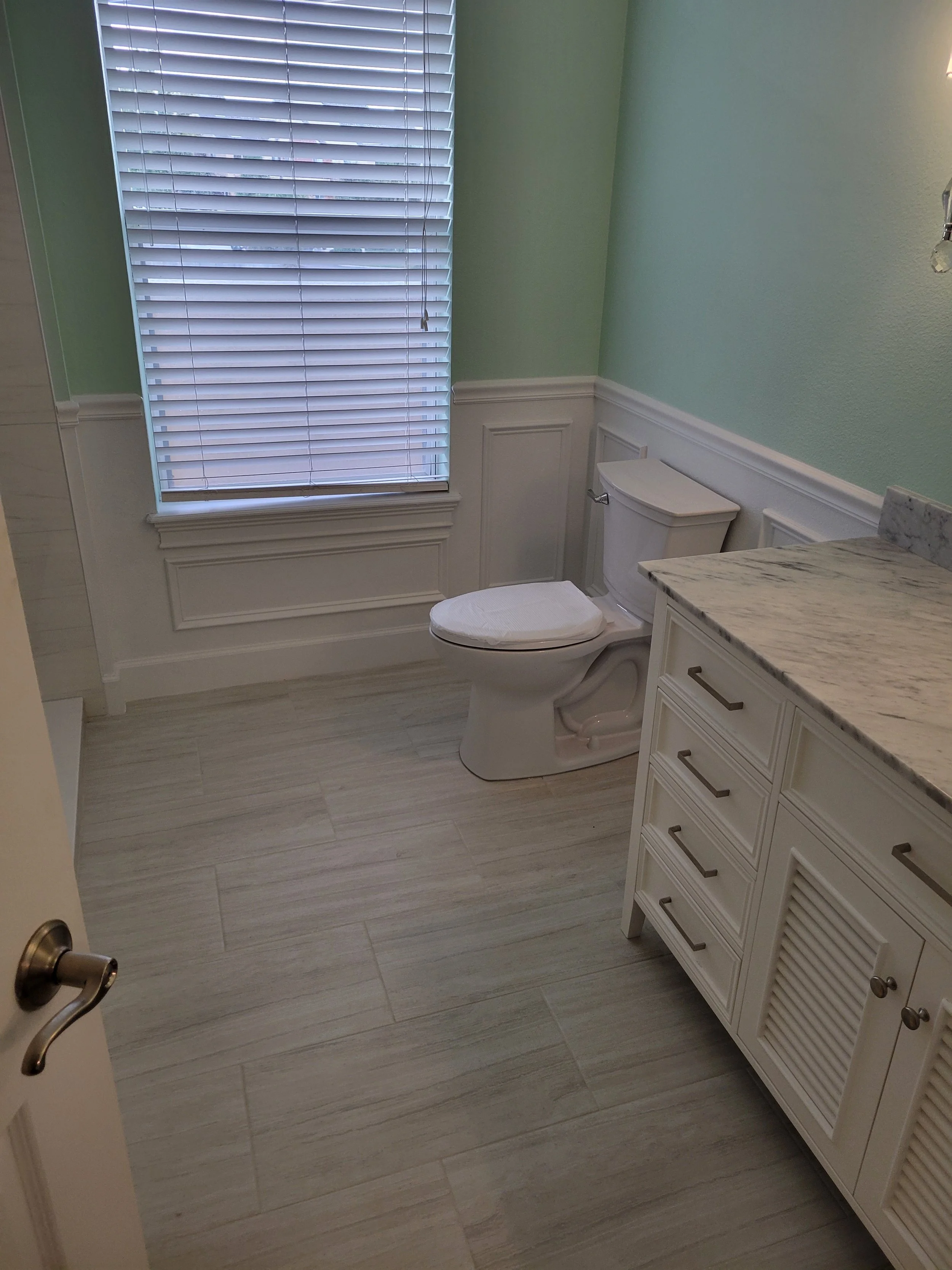 Bathroom with a window with blinds, a white toilet, and a white vanity with a marble countertop.