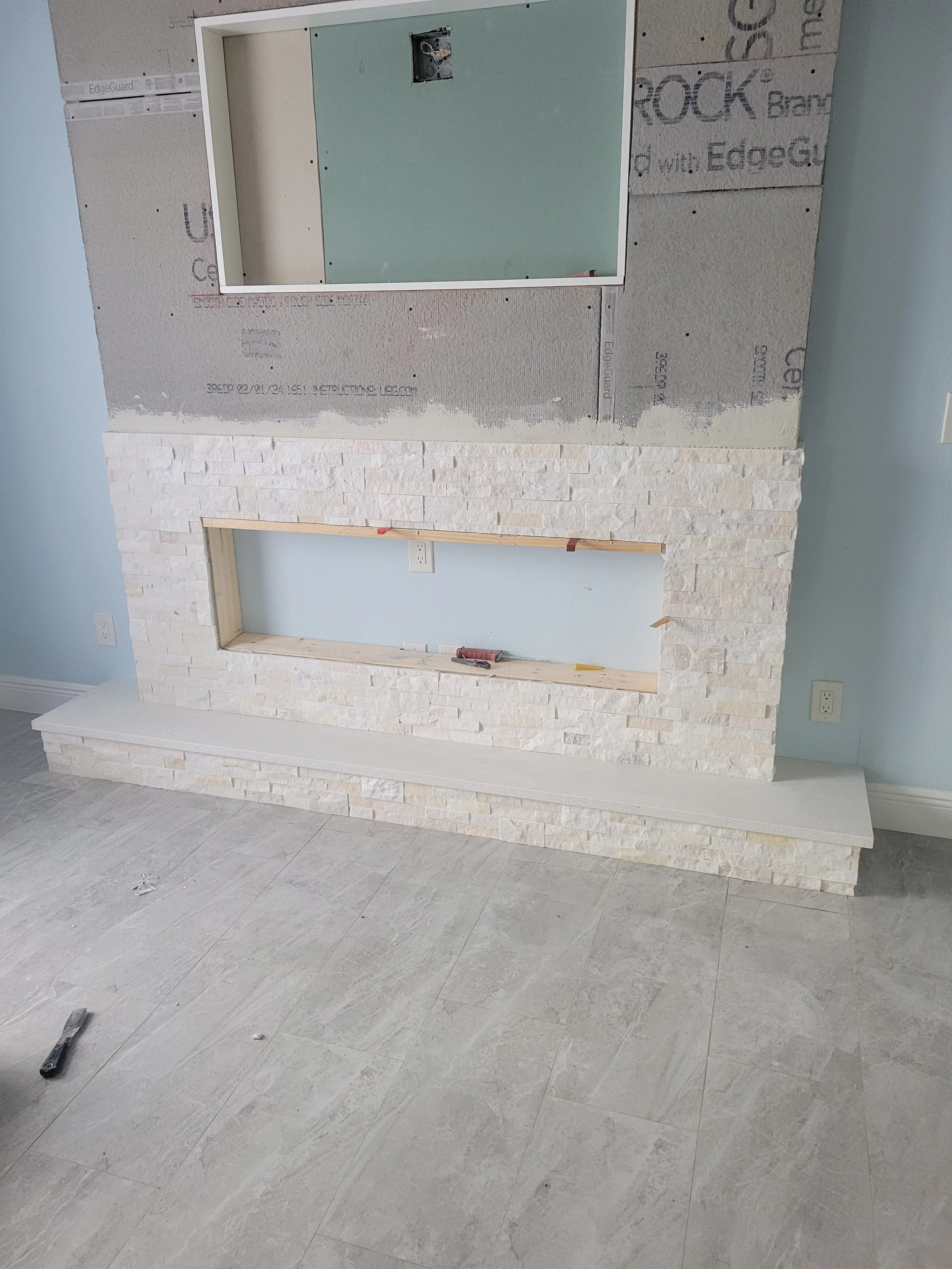 Interior of a room under construction with a partially built fireplace featuring white stone veneer and a wooden mantle, against a light blue wall.