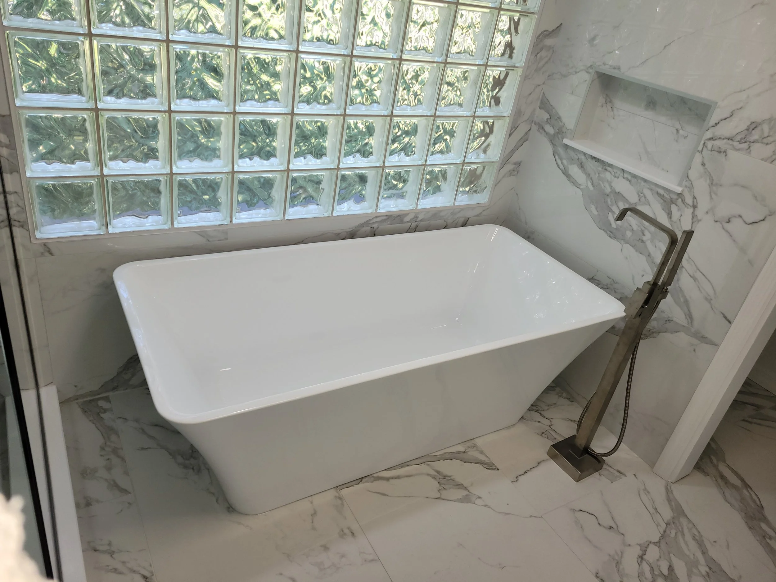 Modern white bathtub with a floor-mounted faucet and a built-in niche, set against marble tiled walls and a glass block window.