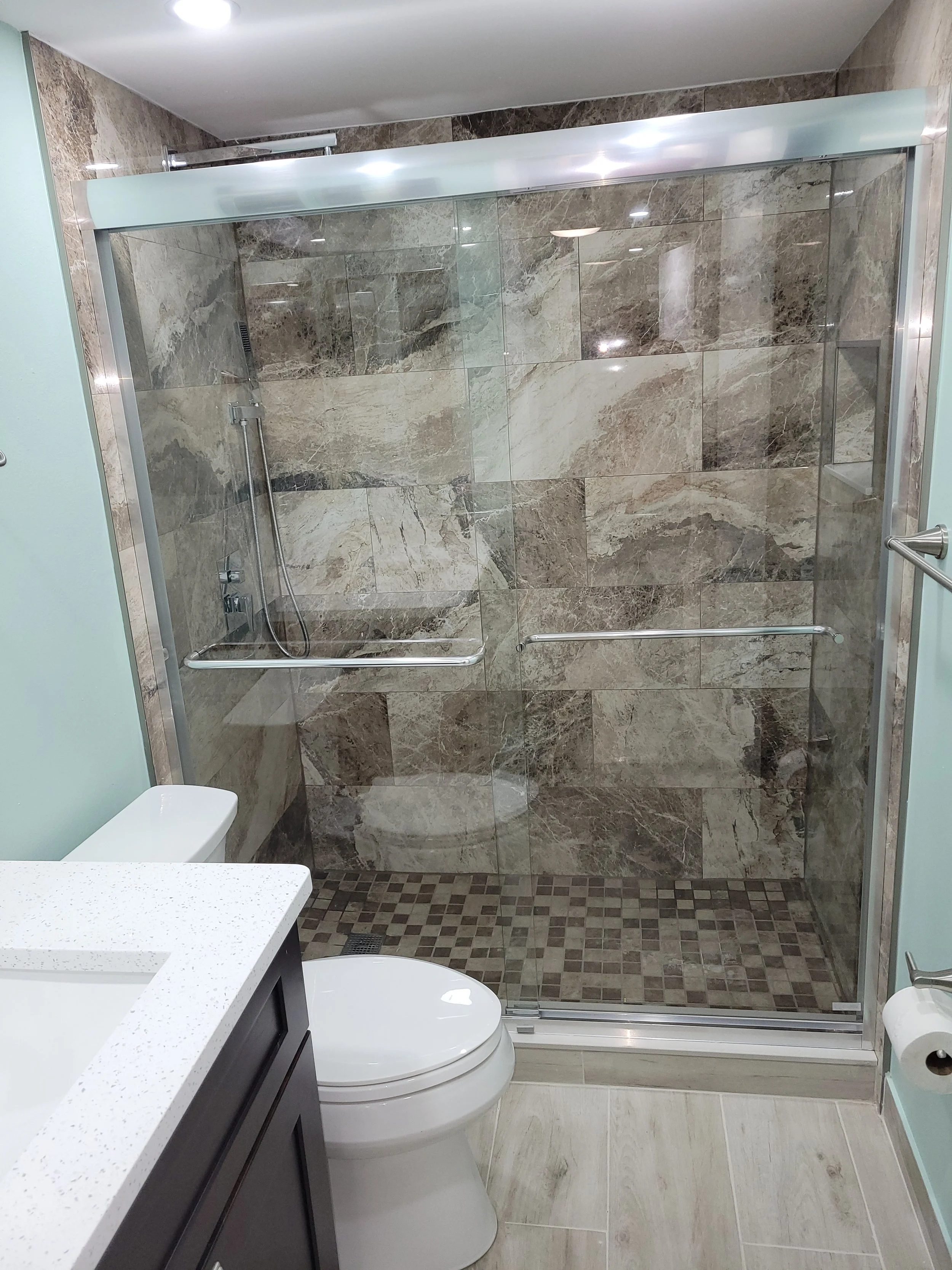 Bathroom with glass-enclosed shower featuring brown marble tiles, a white toilet, a sink with a speckled white countertop, and a toilet paper holder on a light blue wall.
