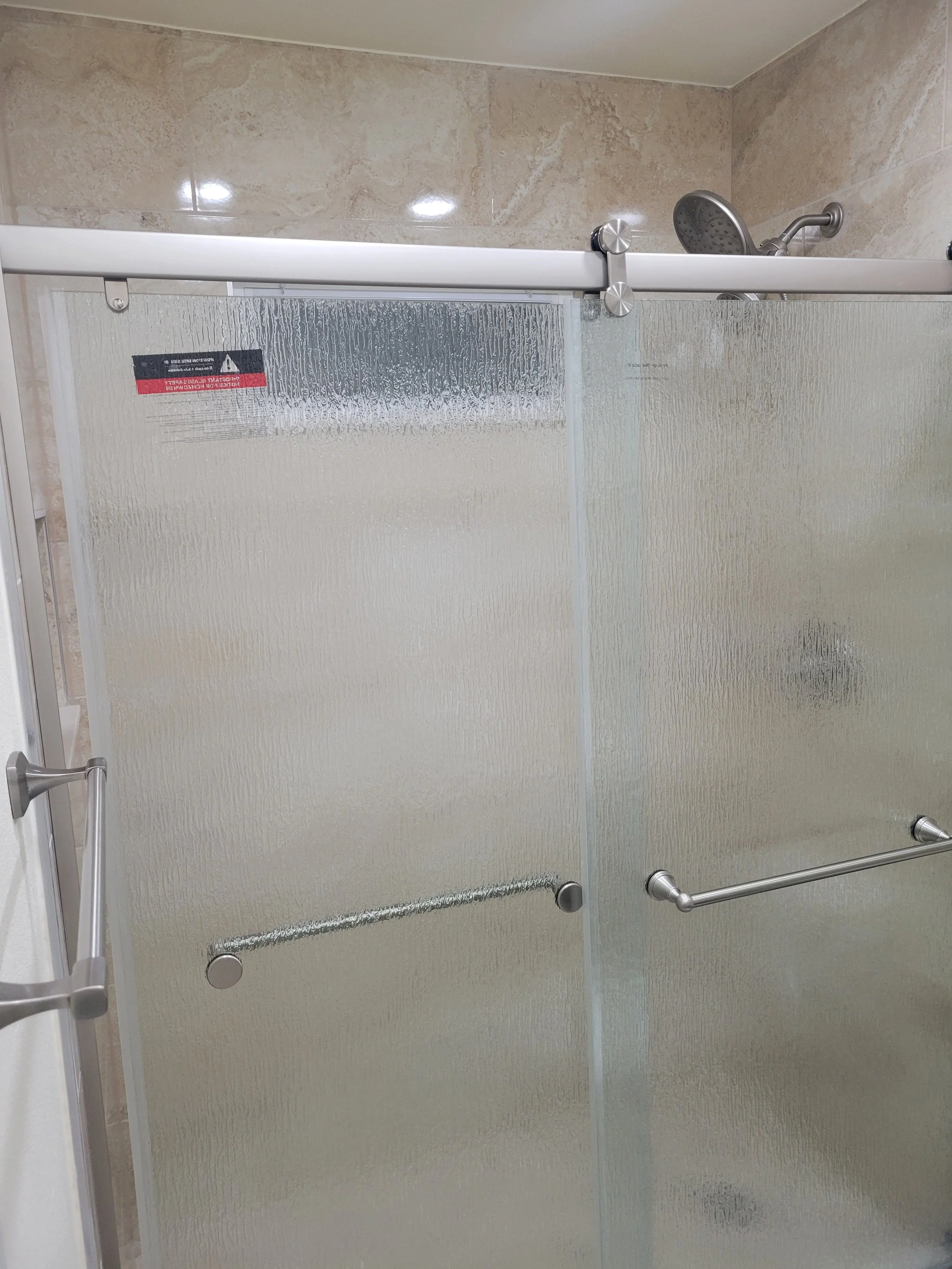 Frosted glass shower doors with metal handles and a sliding door mechanism, showerhead visible inside bathroom with beige tiled walls.
