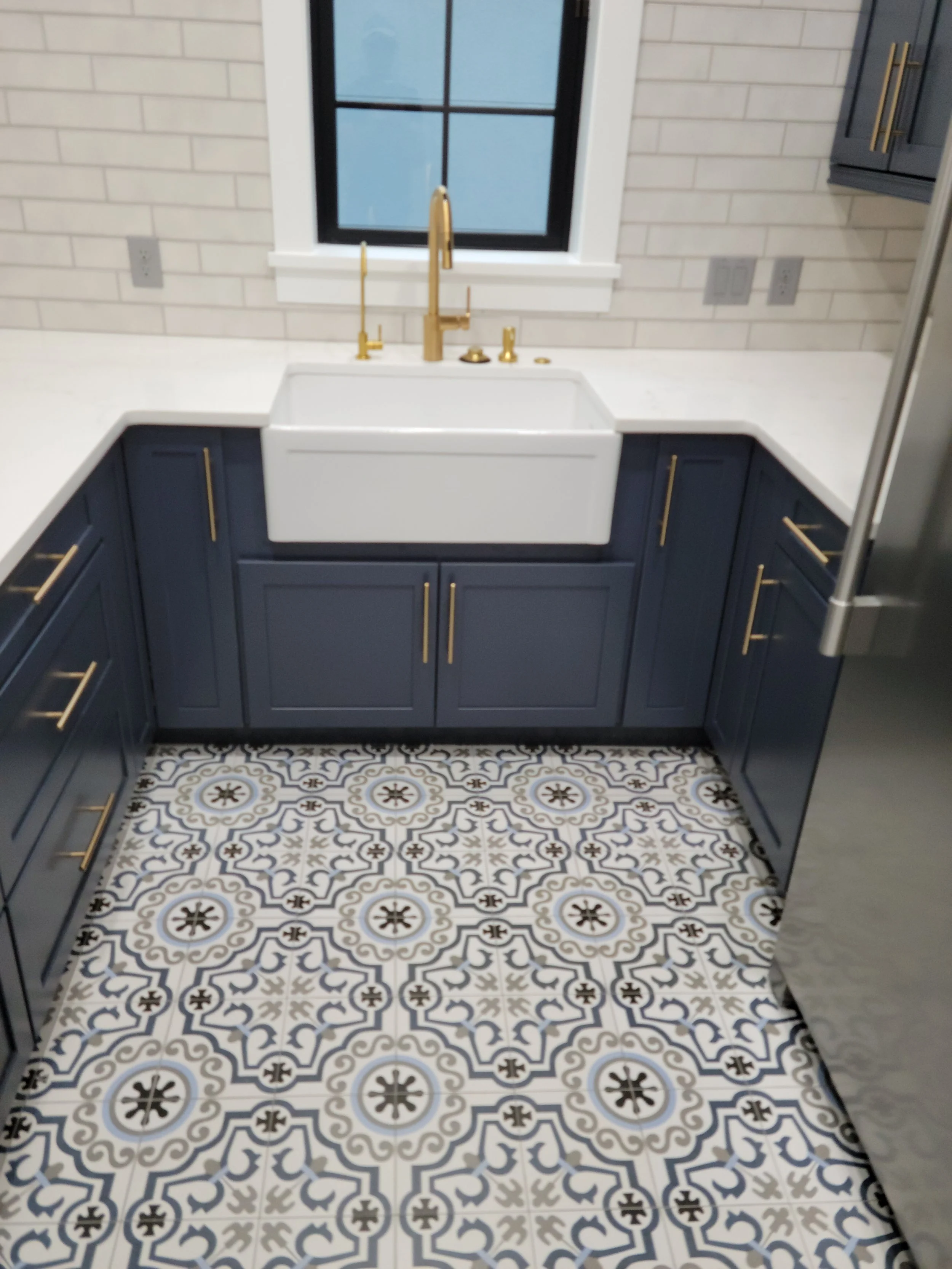 Kitchen with white farmhouse sink, gold faucet, dark blue cabinets with gold handles, patterned tile floor, white brick walls, and a window above the sink.