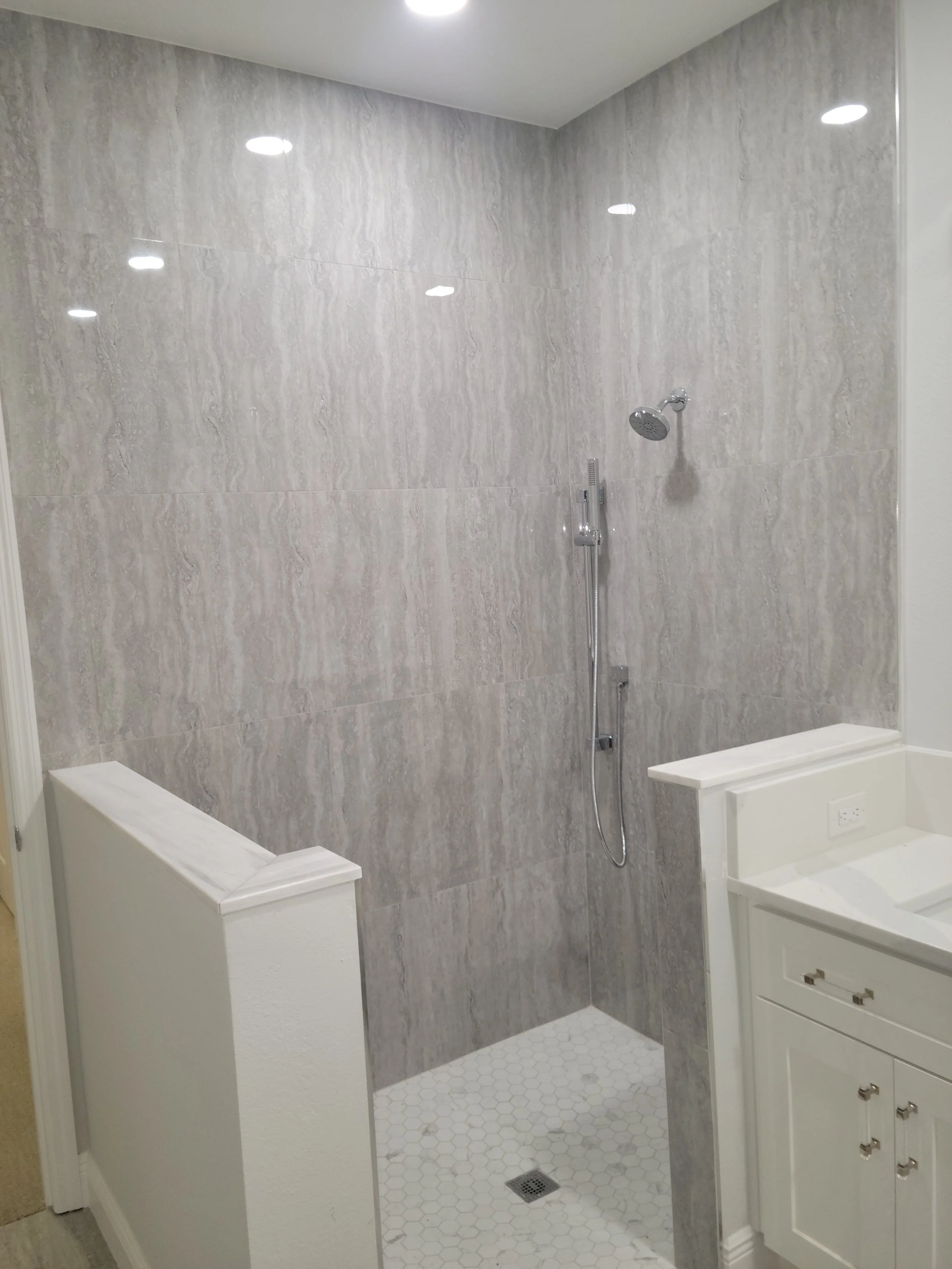 Bathroom with walk-in shower featuring gray stone walls, white hexagonal floor tiles, a silver showerhead, and a white vanity with a single basin.